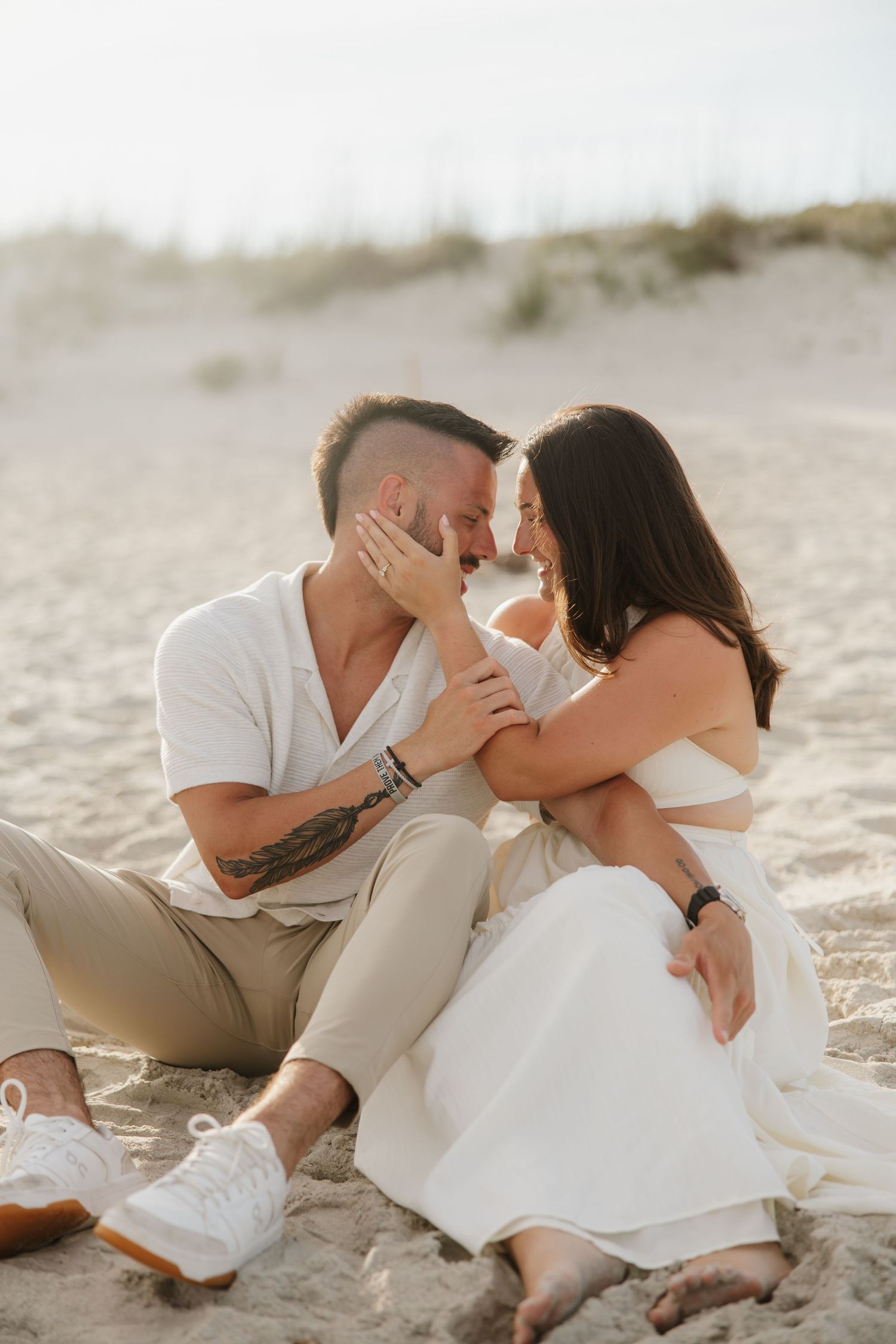 Couple sitting on sandy beach, woman in white dress touching man's face as they lean in for a kiss.