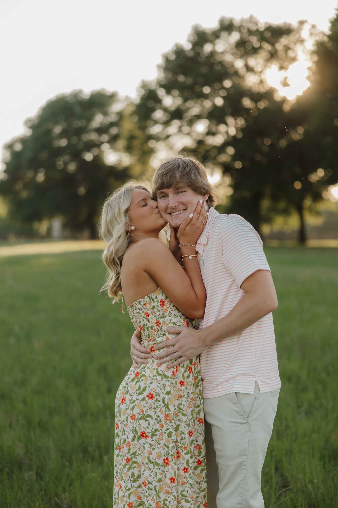 A woman in a floral dress kisses a smiling man on the cheek during a golden hour outdoor photo session.