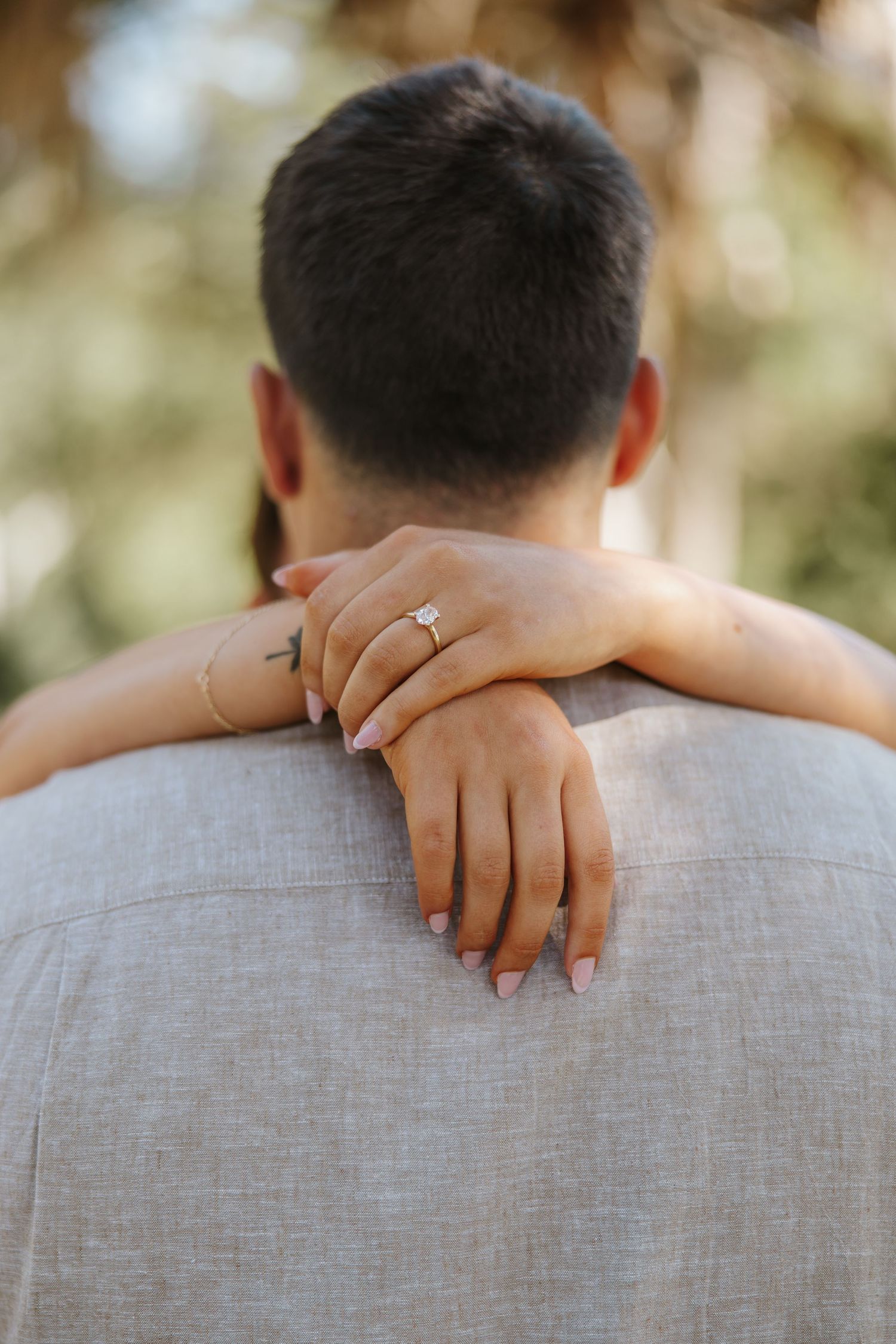 A woman's hand with an engagement ring rests on a man's shoulder from behind, set against a blurred outdoor background.
