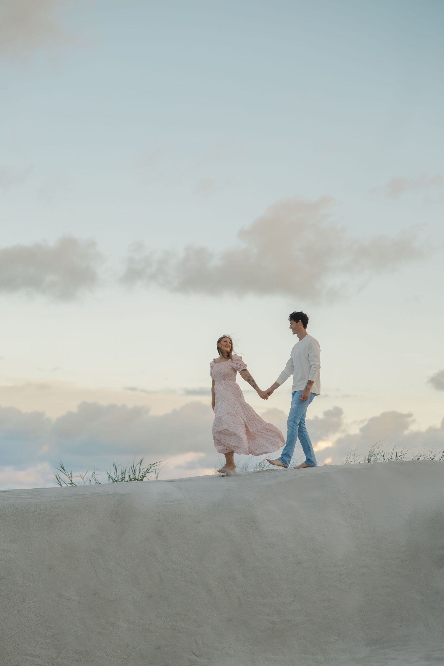 Couple holding hands walking on sand dunes at sunset, woman in pink dress, man in white shirt and jeans.