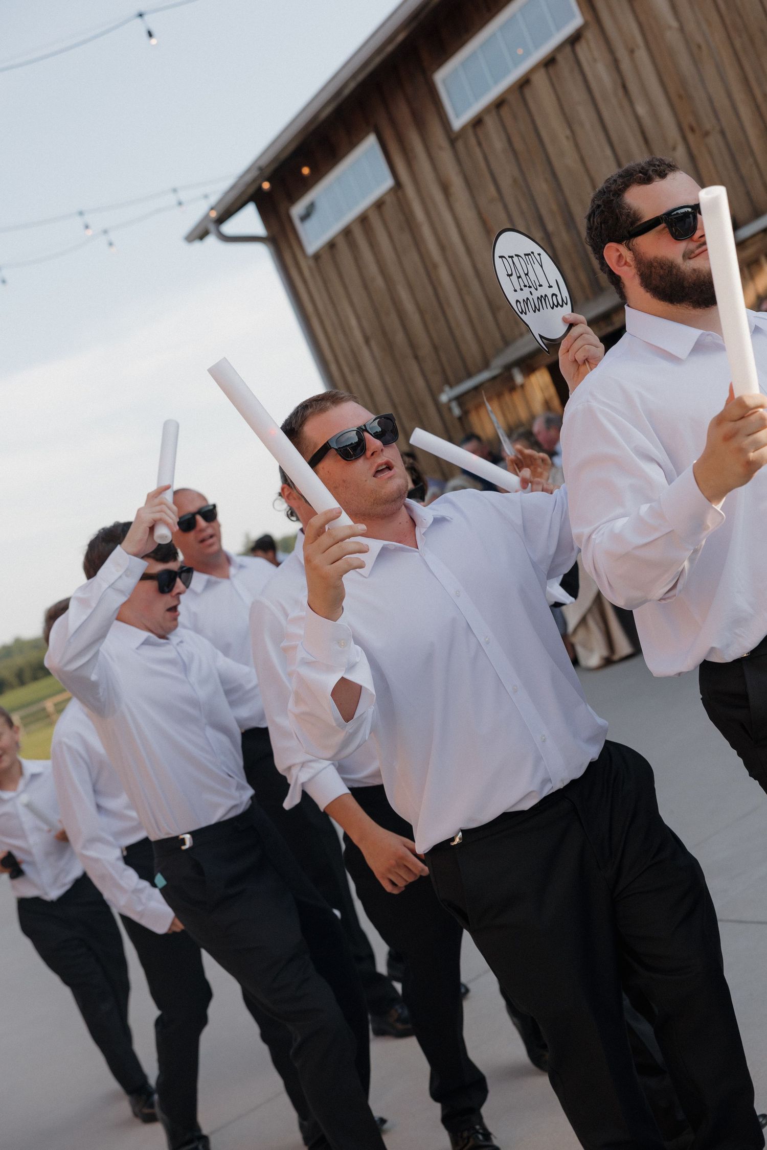 Groomsmen in white shirts, black pants and sunglasses dance with glow sticks outside a rustic barn at a wedding reception.