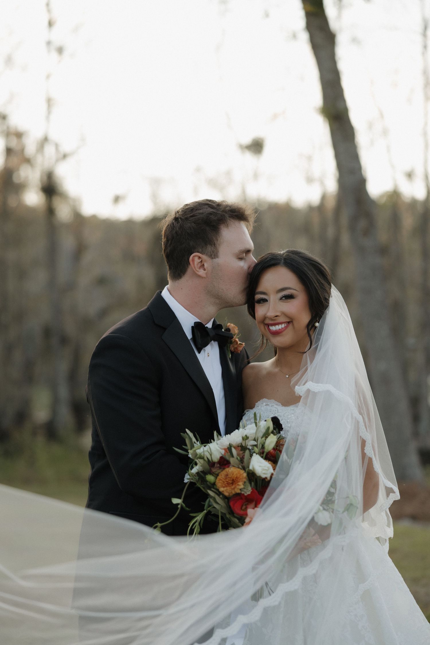 Groom in a black tuxedo kisses his smiling bride's forehead as she holds an autumn bouquet, her long veil flowing in a woodland setting.