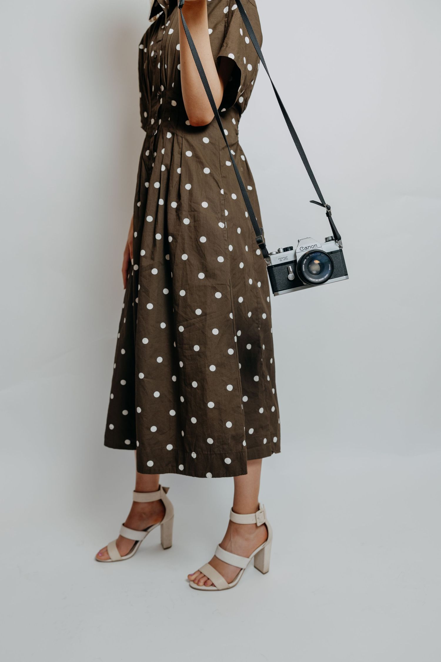 Woman in an olive polka dot midi dress with a vintage film camera and white block-heel sandals.