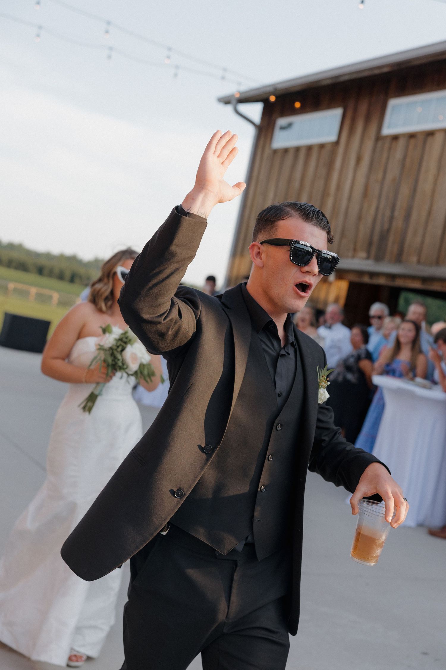Groomsman in black suit and sunglasses raises his hand while holding a beer at an outdoor rustic barn wedding reception with guests nearby.
