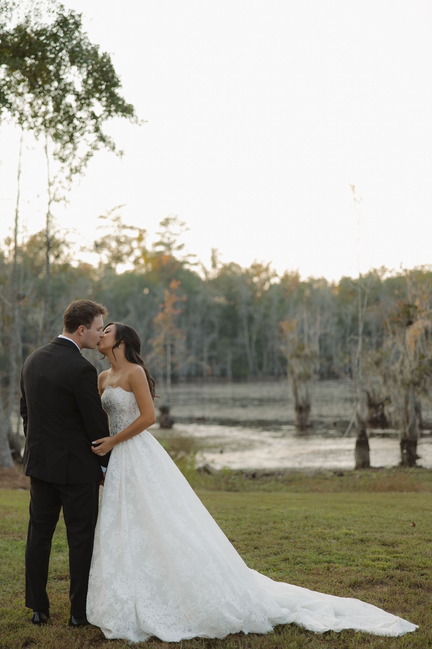 Bride in a strapless lace wedding gown and groom in a black suit share a kiss beside a misty cypress swamp at sunset.