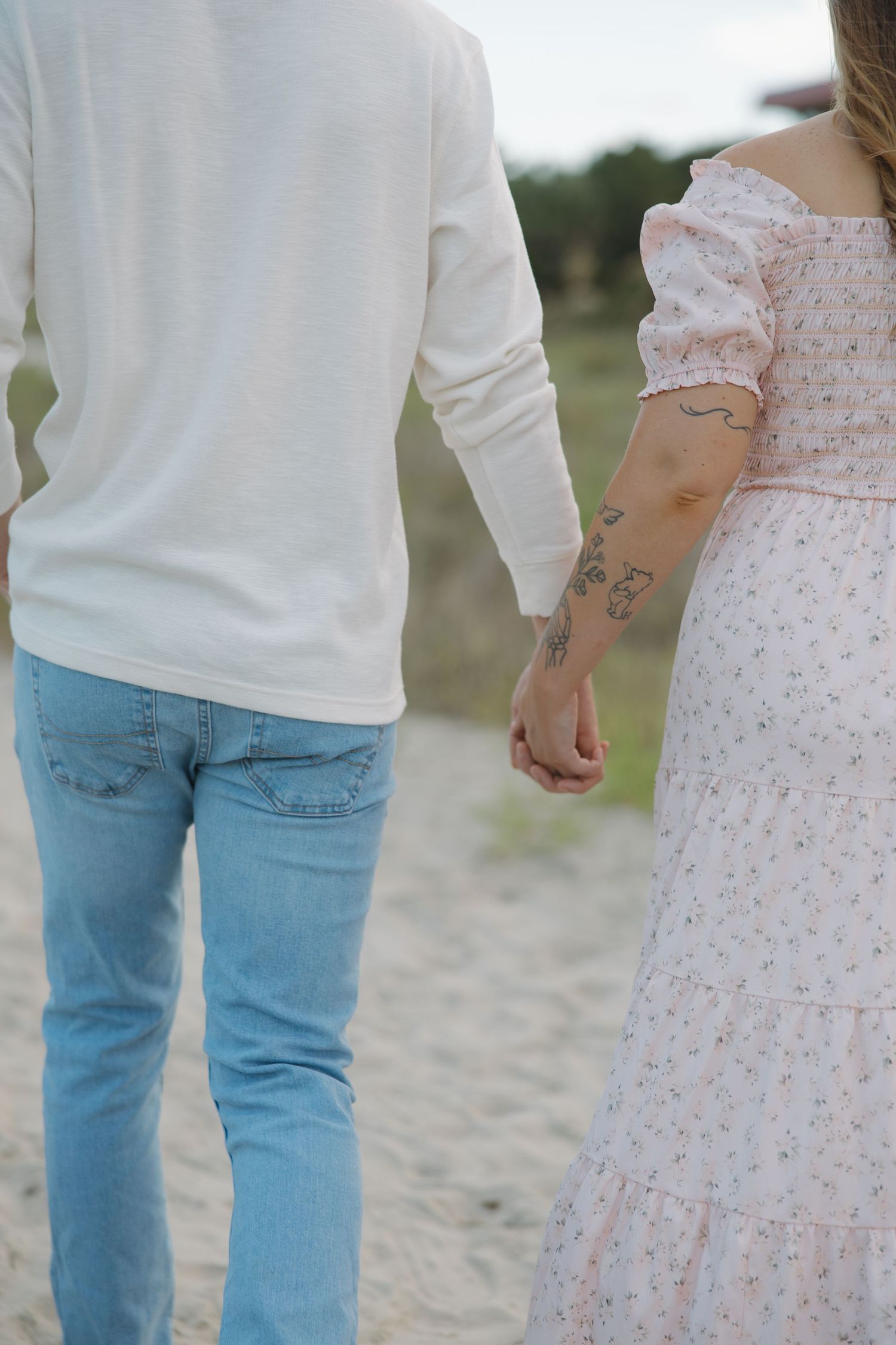 Couple holding hands on sandy beach path, woman wearing floral dress with tattoos, man in white shirt and jeans.