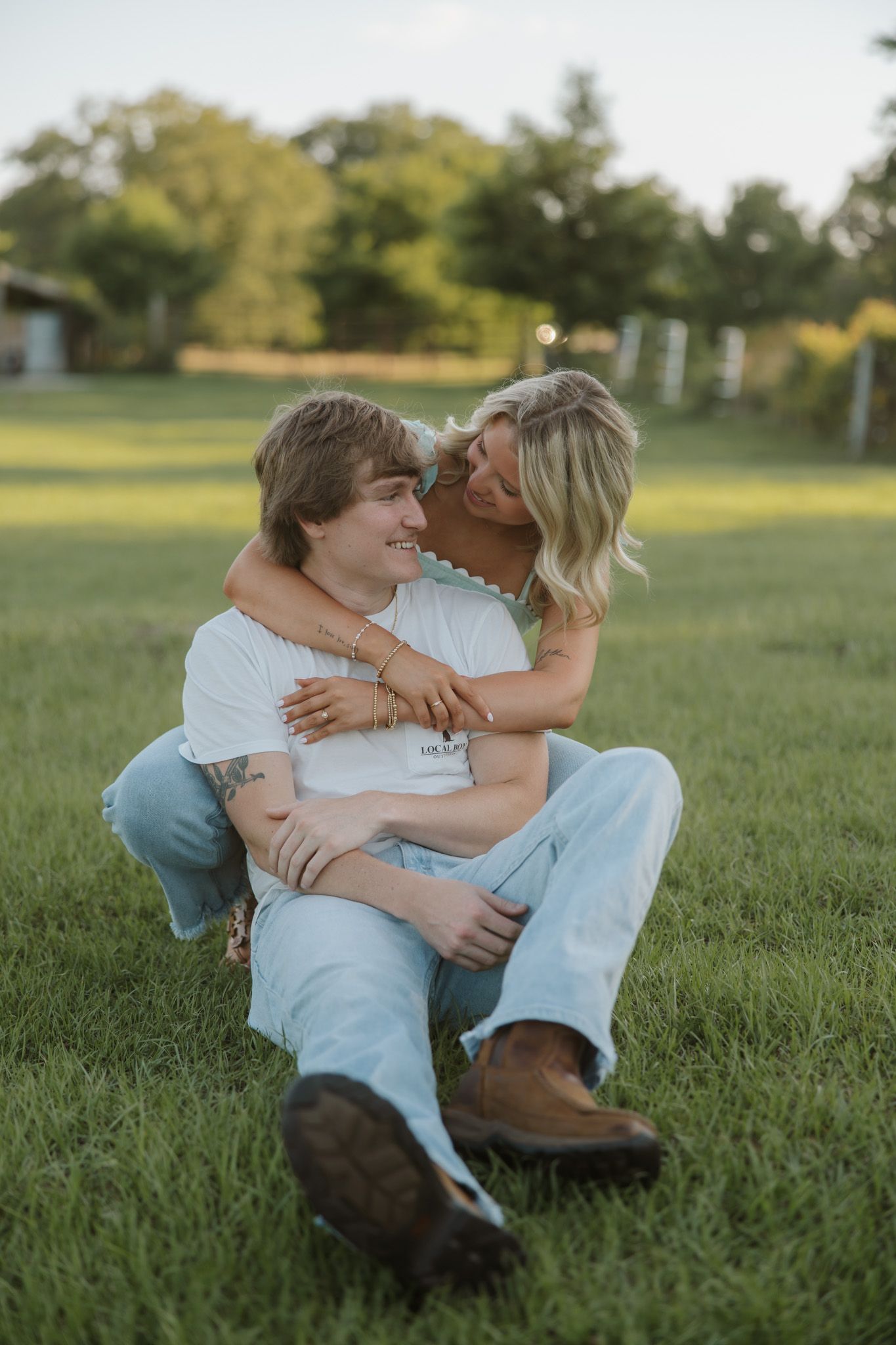 A young couple sitting on green grass, embracing and smiling, with trees and a fence in the background.