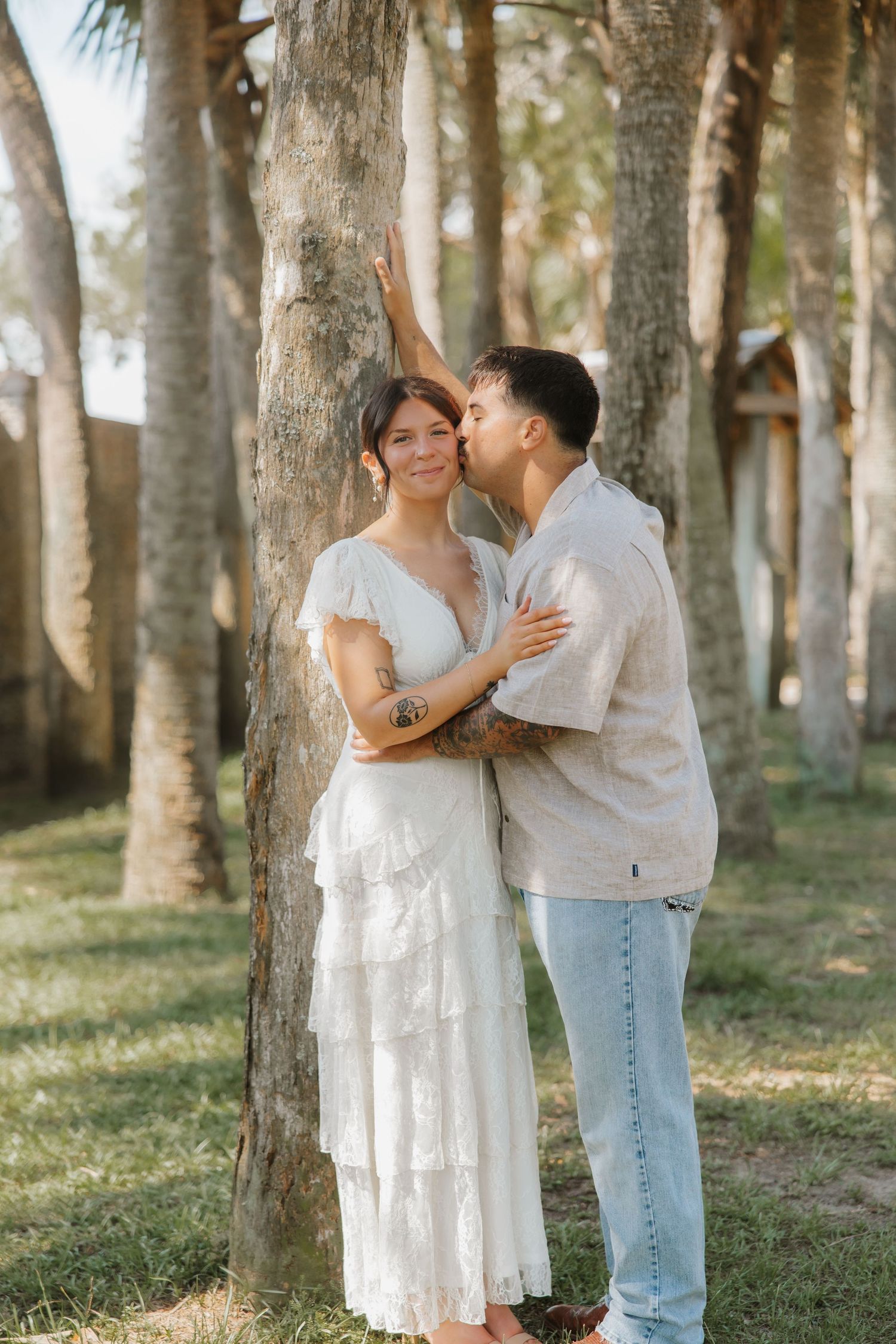 A couple shares a romantic moment among palm trees, man kissing woman's cheek as she smiles in a white dress.