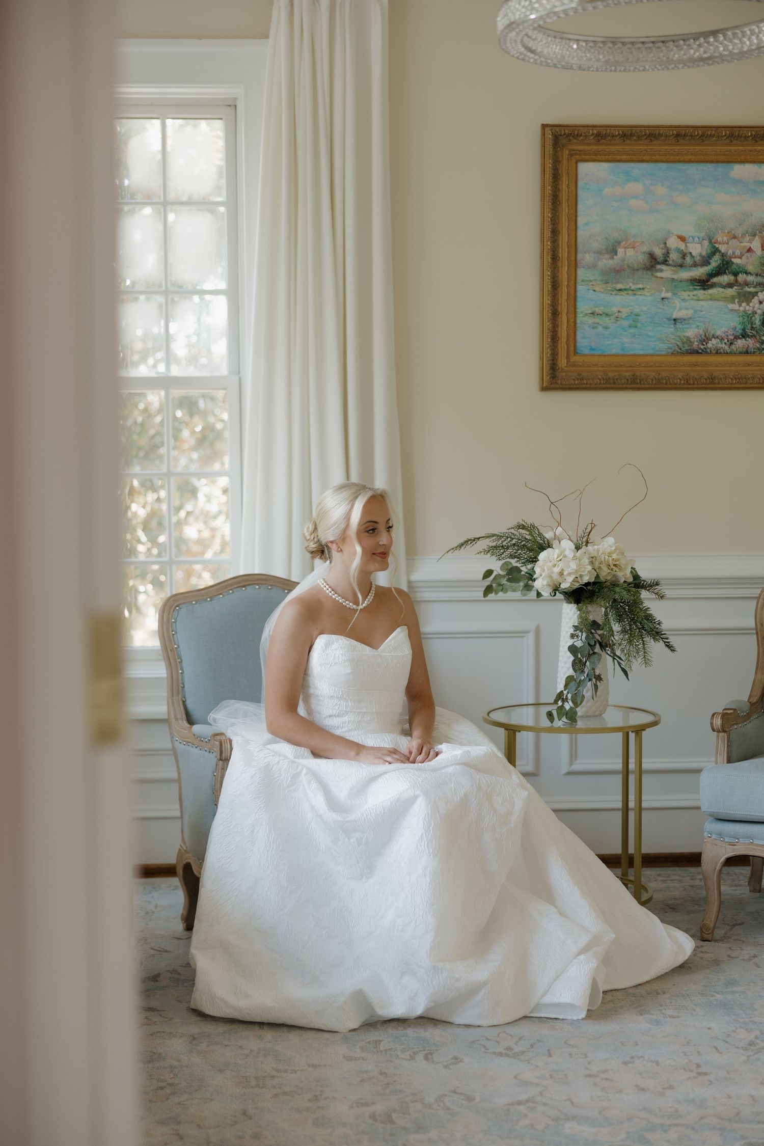 Elegant bride in a white strapless gown seated in a blue French chair near a sunlit window in a classic interior.