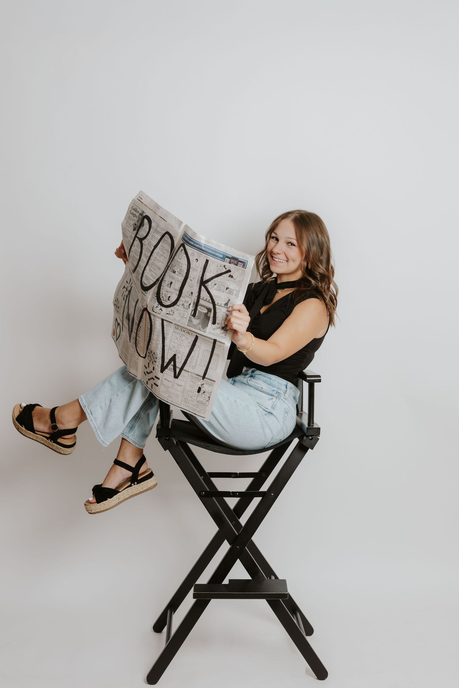 Smiling woman sits in director chair holding a Book Now newspaper sign on white background.