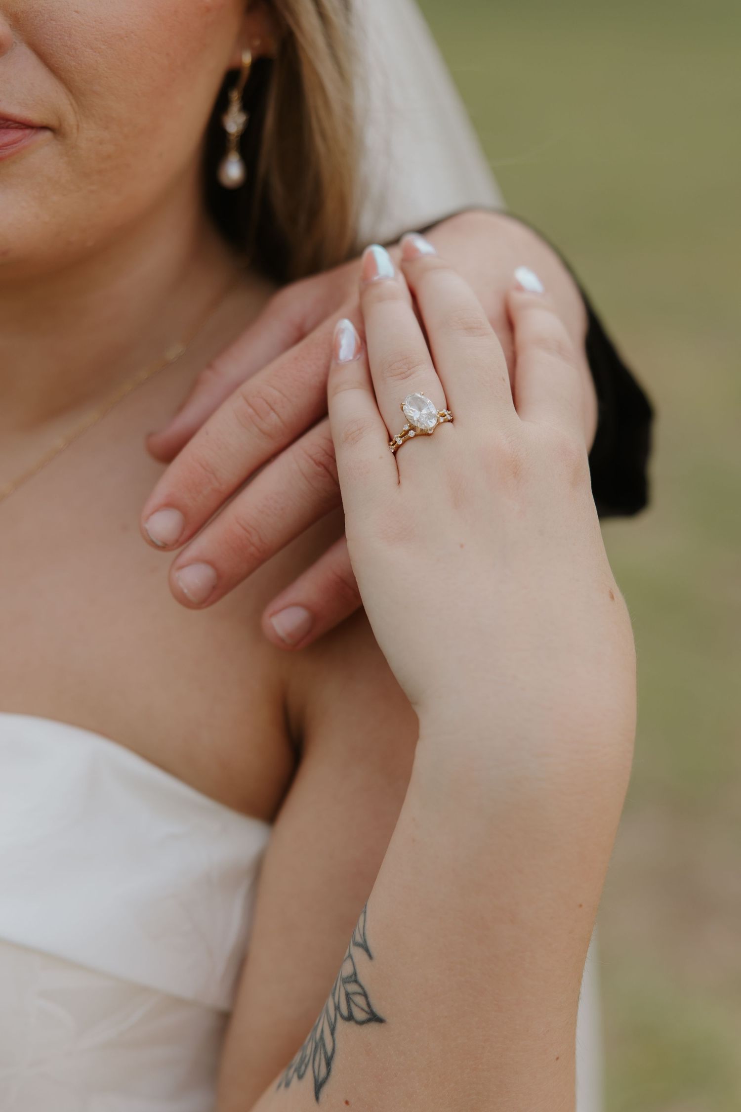 Close-up of bride's hand displaying a gold pear-shaped engagement ring, with pearl earring and tattoo visible.