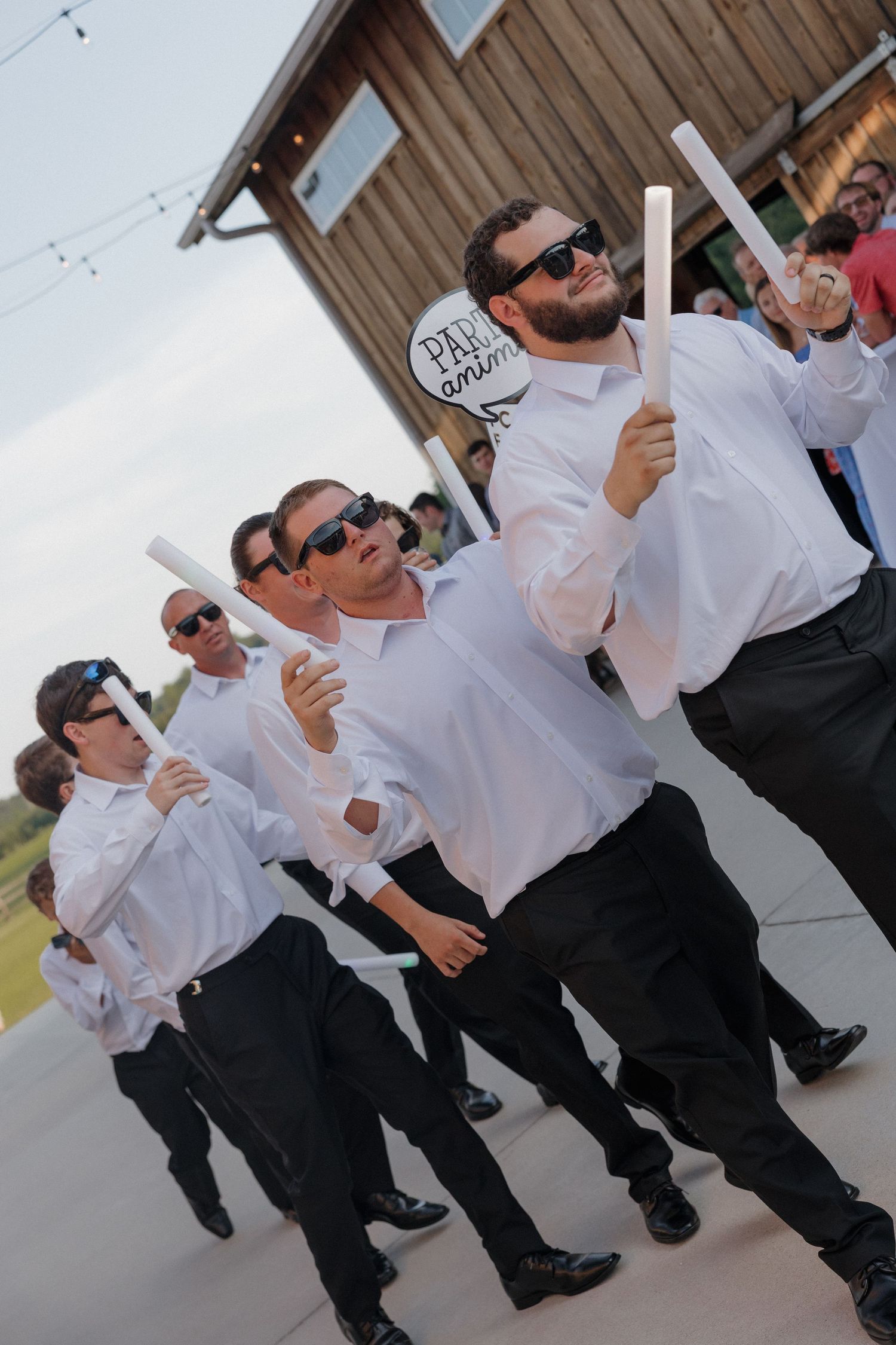 Groomsmen in white shirts, black pants and sunglasses walk in a procession carrying white candles outside a rustic wooden venue.
