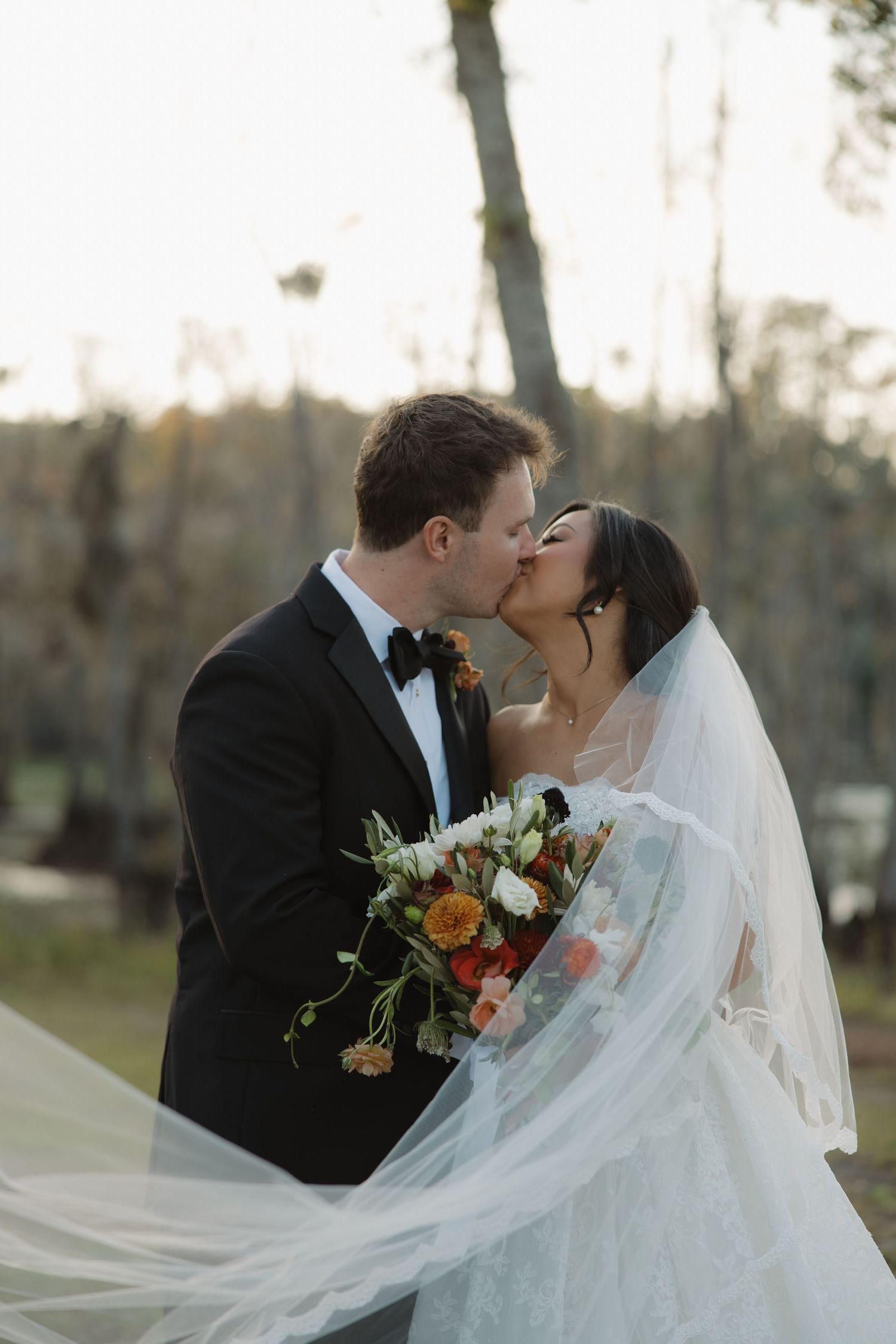 A groom in a black tuxedo kisses his bride in a flowing veil as she holds an autumn bouquet outdoors.