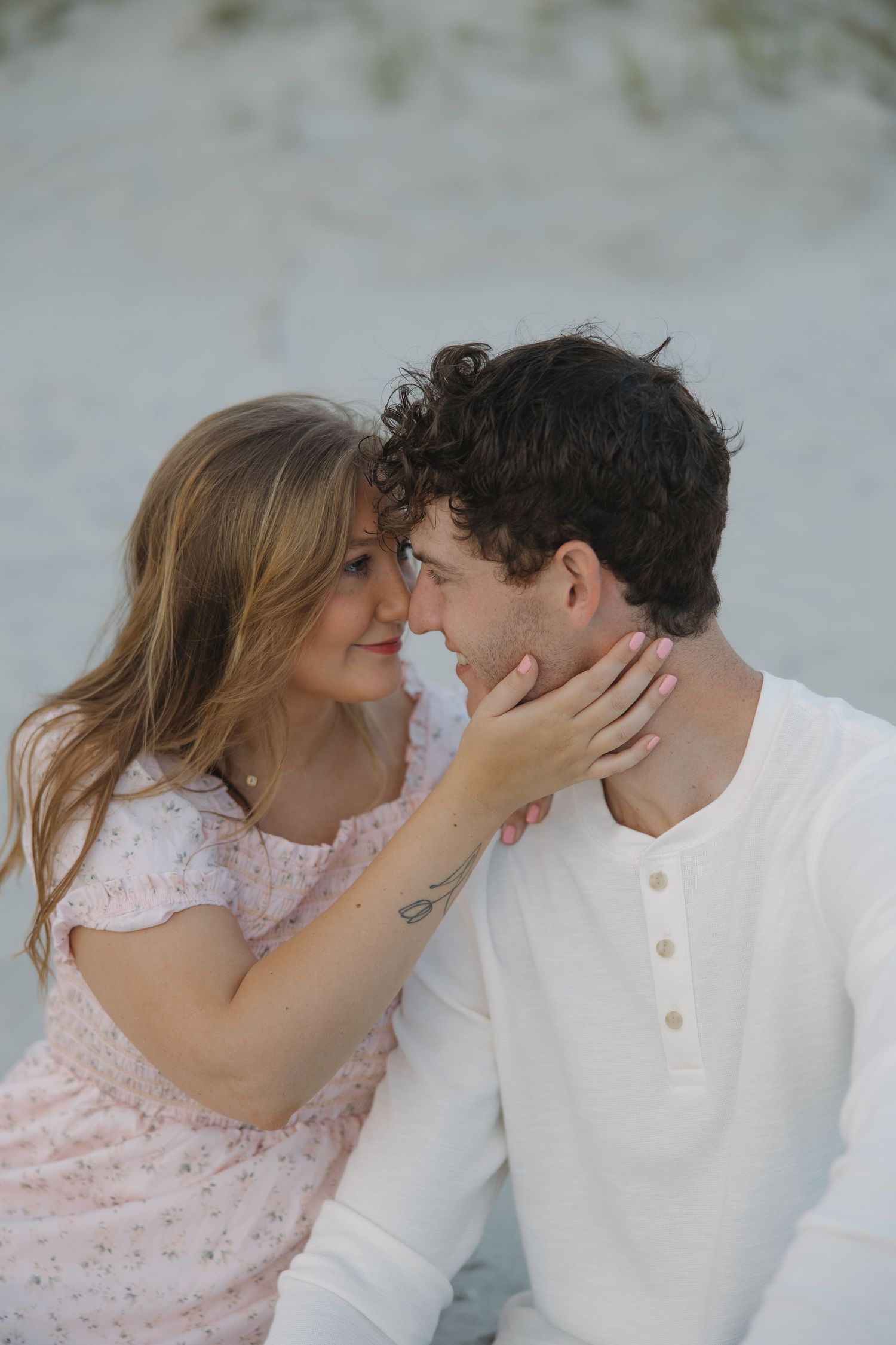 Couple sharing a romantic moment on a sandy beach, woman in pink floral dress touching mans face tenderly.