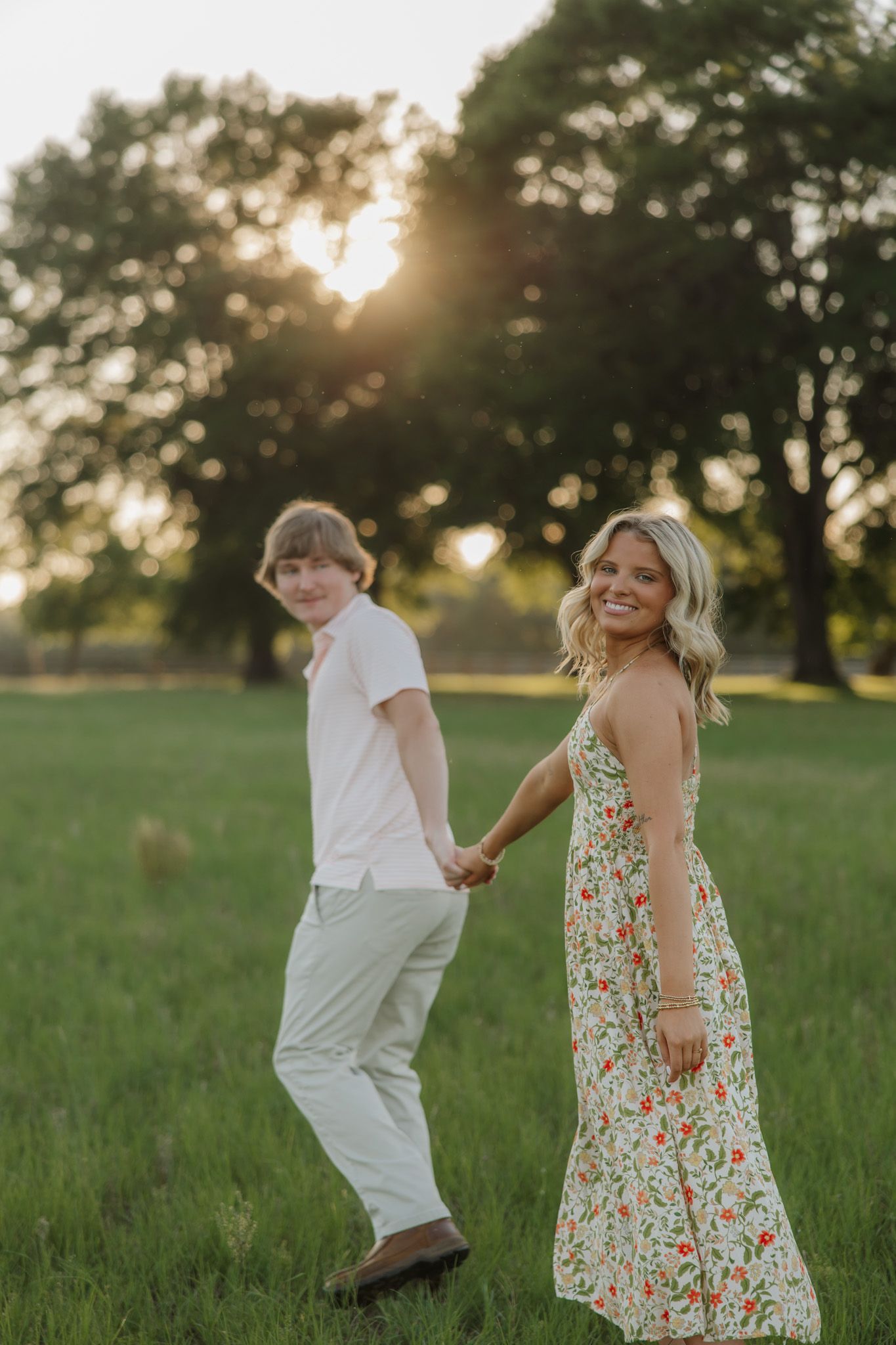 A young couple holds hands smiling in a sunlit green field with trees at golden hour.