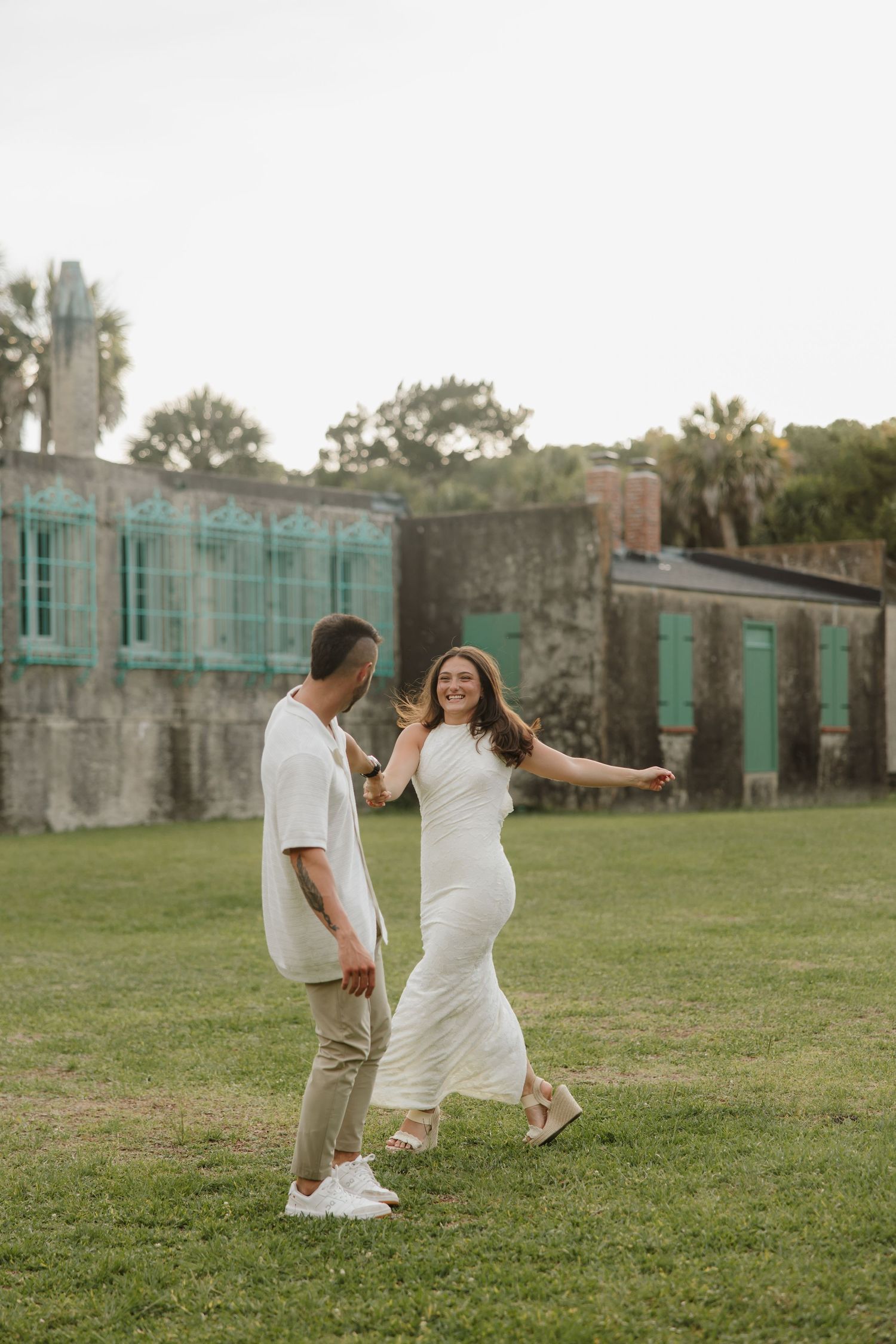A couple dances joyfully on green grass near historic ruins with teal windows and palm trees.