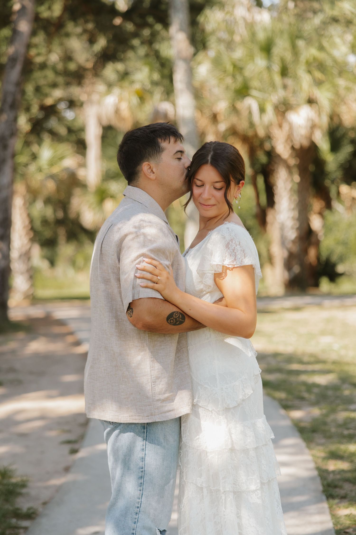 A couple shares a tender moment outdoors, man kissing woman's temple, both in casual white attire among palm trees.