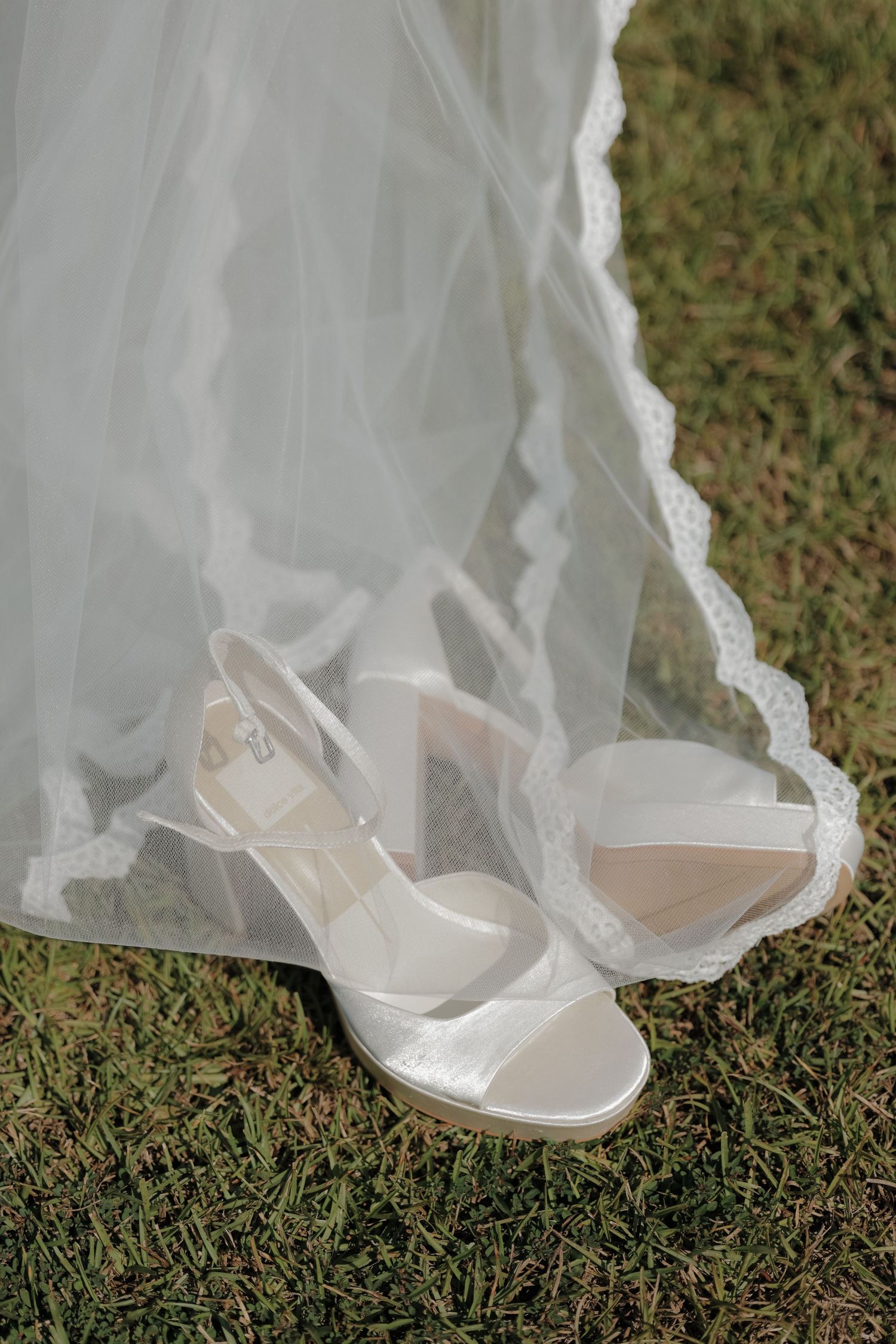 White satin platform bridal sandals with ankle straps resting on green grass beneath a sheer lace-trimmed wedding veil.