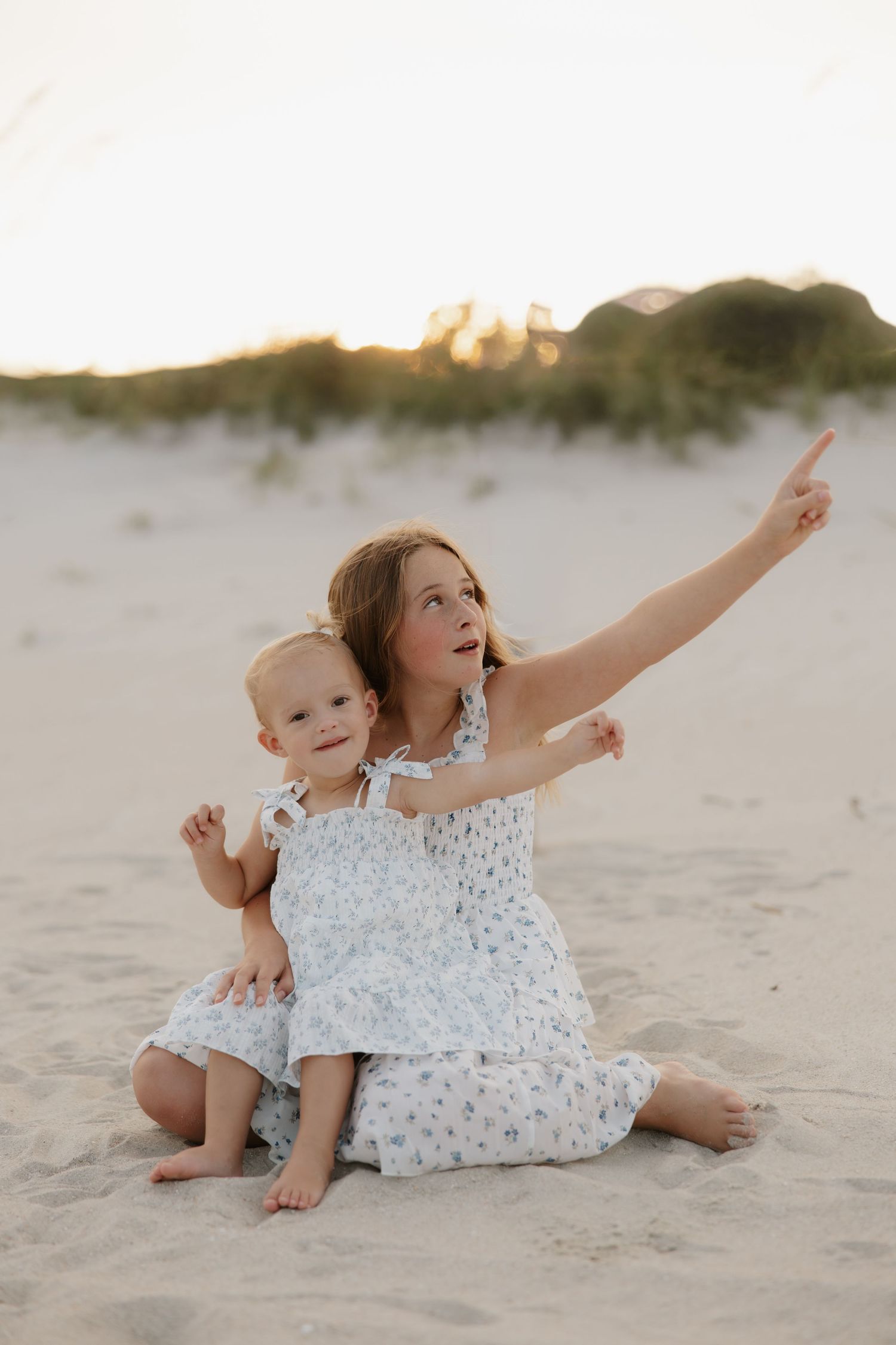 Two young girls in white floral dresses sit on sandy beach at sunset, older sister pointing at the sky.