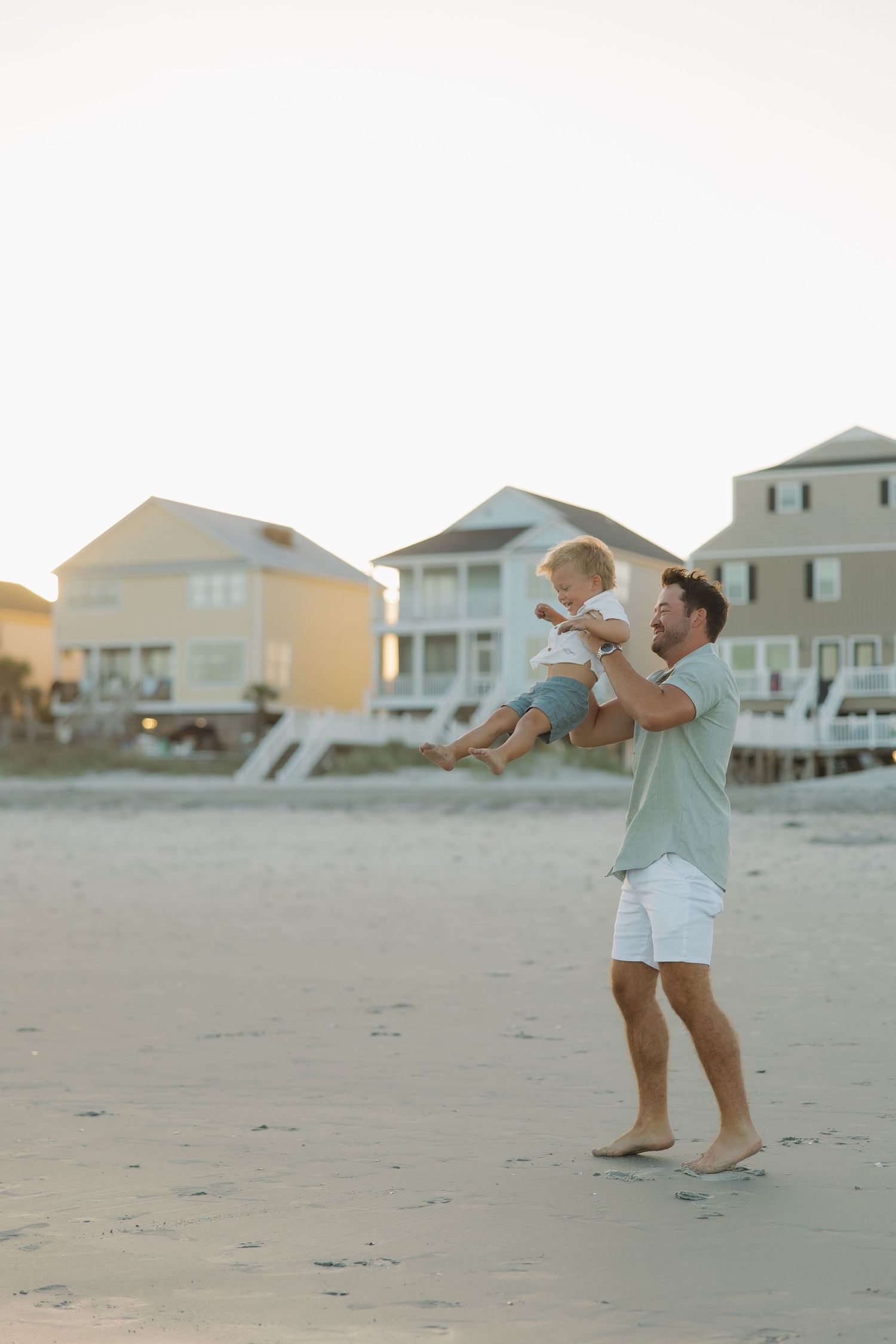 Father joyfully lifting toddler on a sandy beach at dusk with coastal vacation homes in the background.