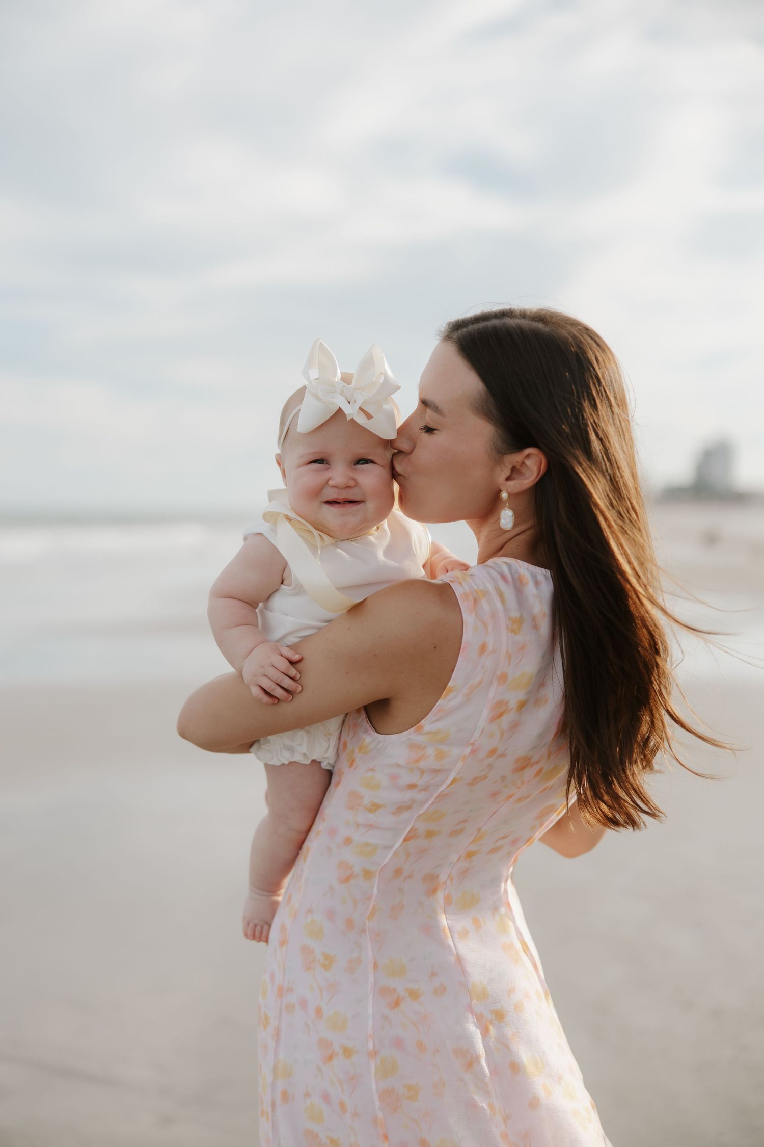A mother in a floral dress kisses her smiling baby girl wearing a white bow on a beach.