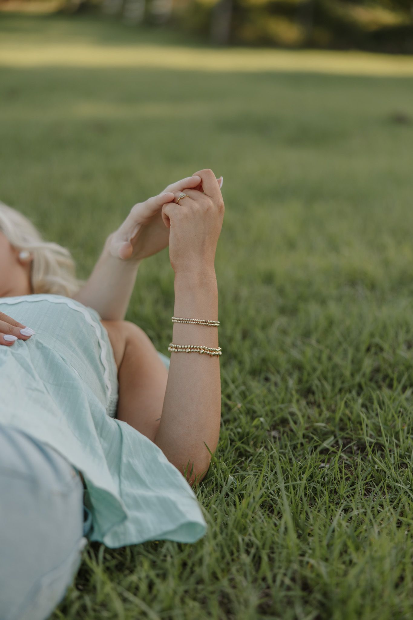 Woman lying on green grass wearing stacked gold bracelets, hands clasped together in soft natural light.