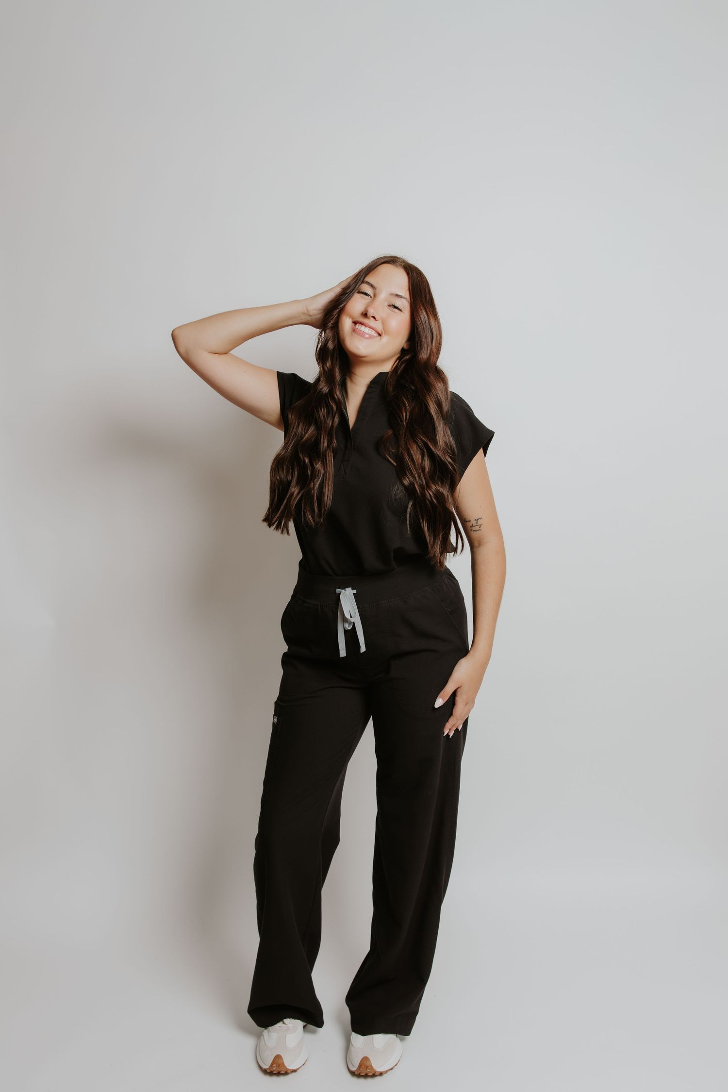 Woman in black jumpsuit with drawstring waist and white sneakers, smiling with hand in hair against white background.