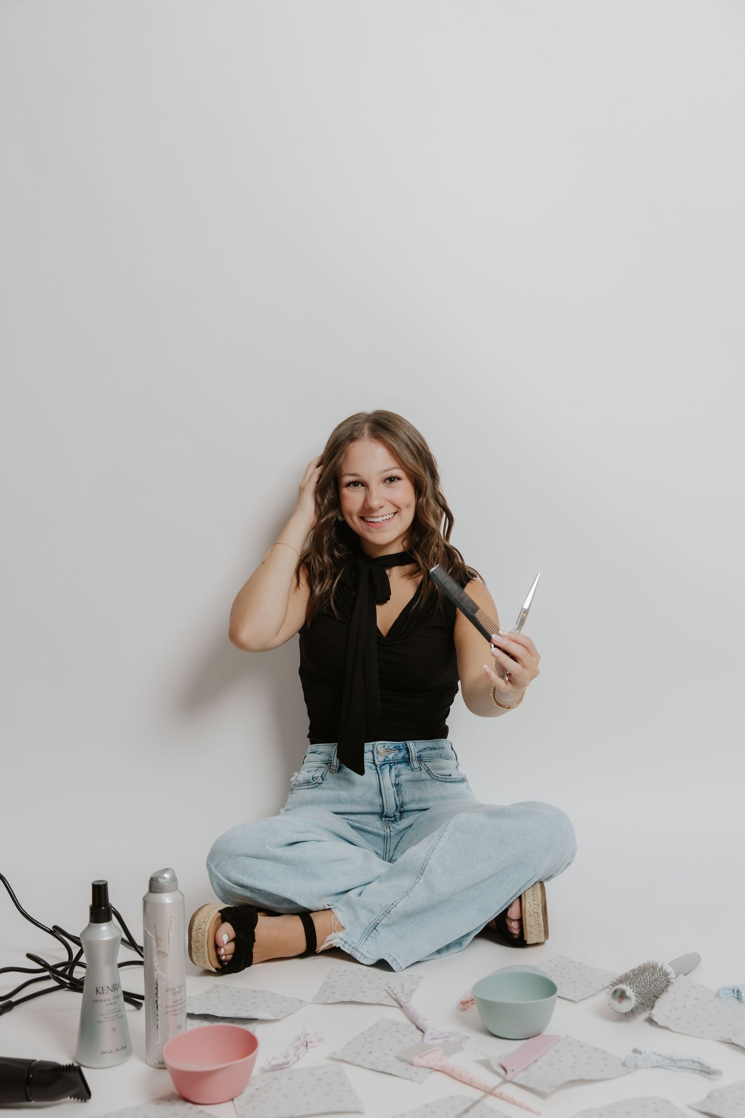 Female hairstylist sitting on floor surrounded by hair tools holding scissors in black top and jeans.