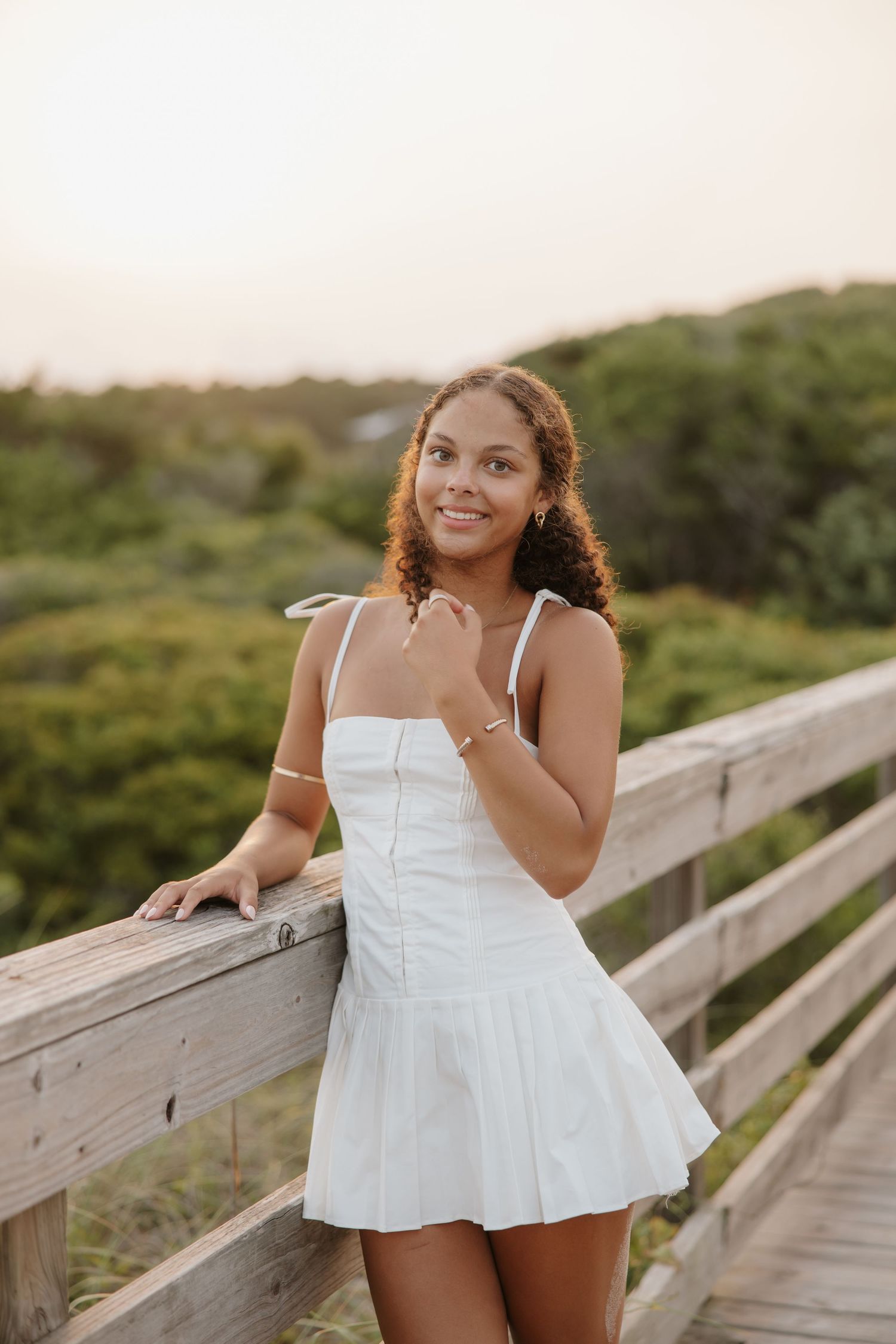 Young woman in white dress smiling on wooden boardwalk surrounded by lush greenery at sunset.