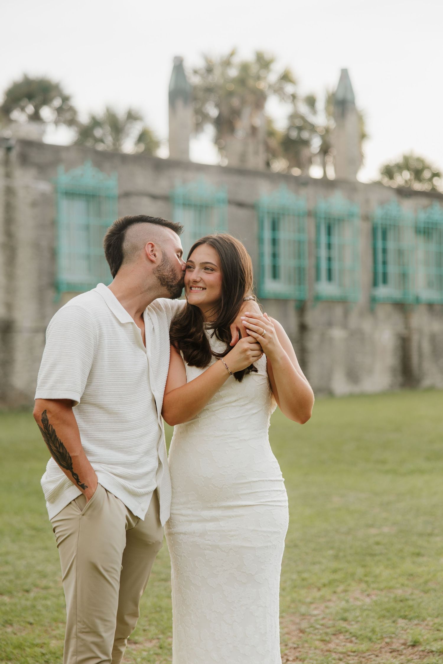 Couple poses outdoors near historic stone building, man kisses pregnant woman's cheek as she smiles warmly.