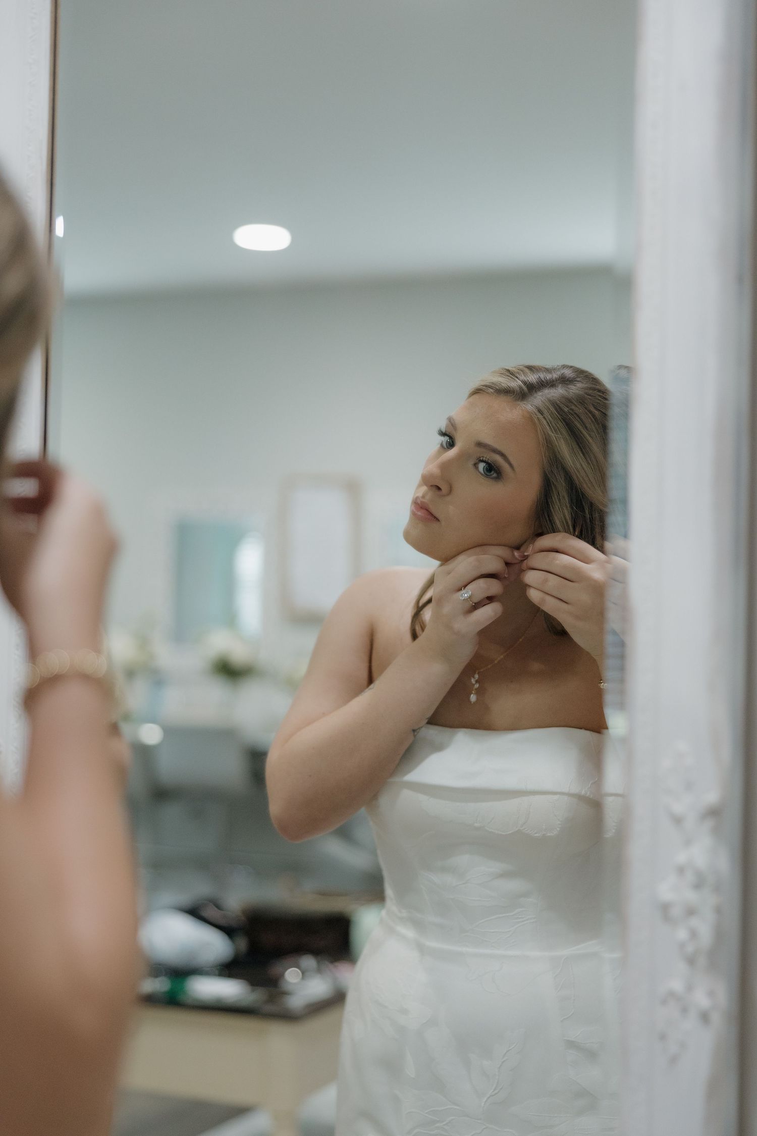 Bride in a white strapless wedding dress adjusts her earring while looking at her reflection in a white ornate mirror.