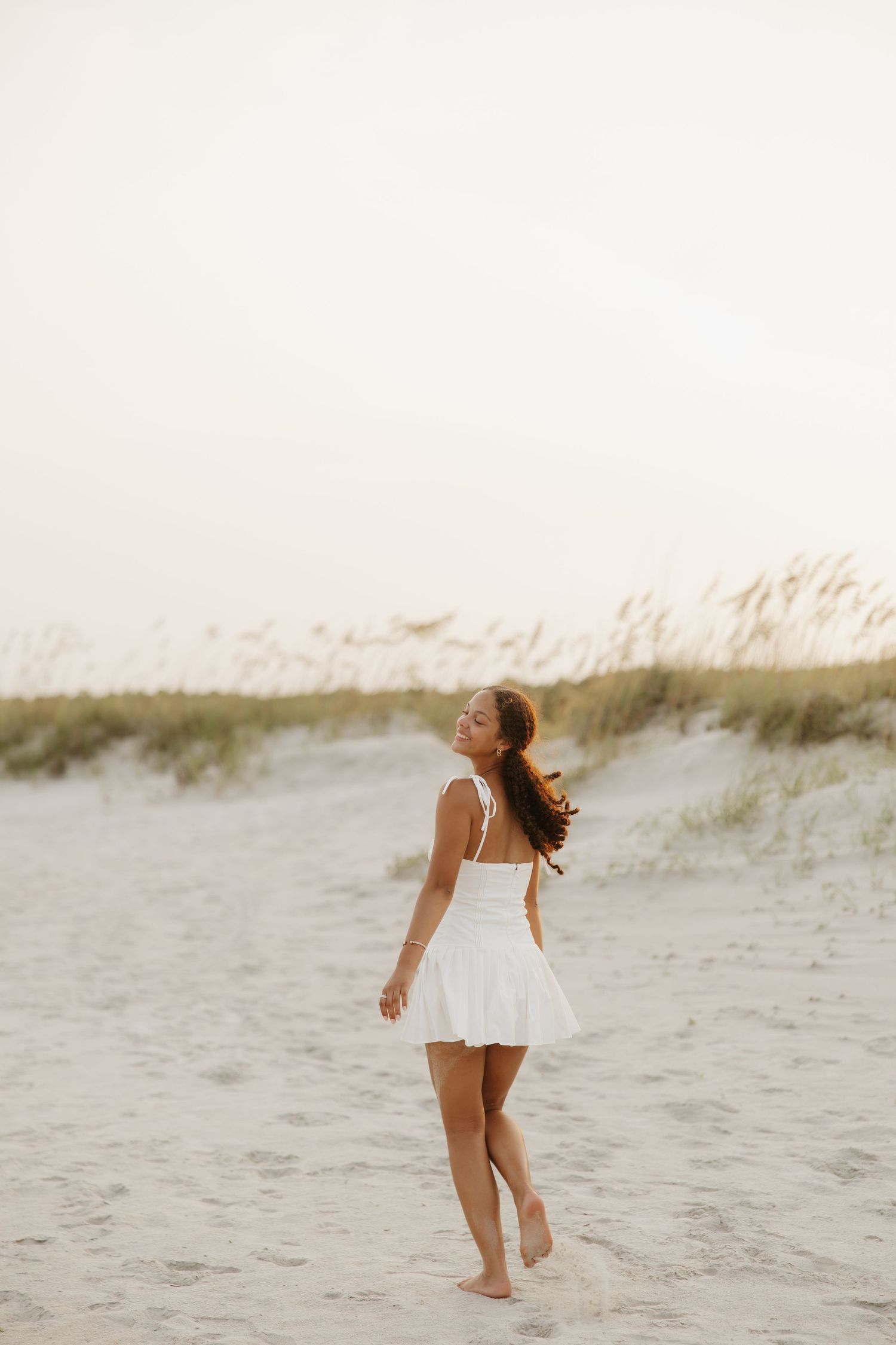 Young woman in white dress walking barefoot on sandy beach dunes at sunset, hair flowing in the breeze.