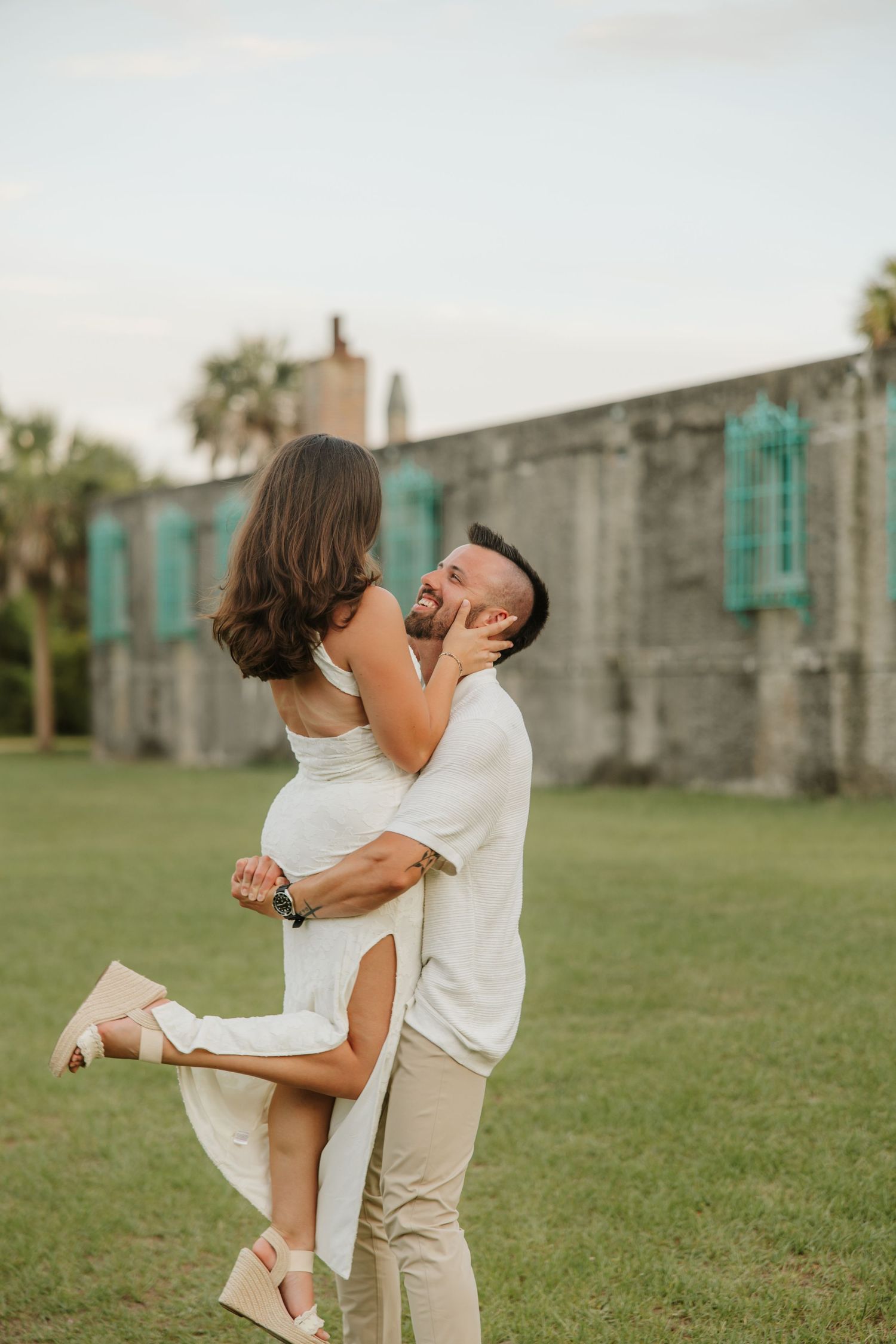 A joyful couple embraces outdoors, man lifting woman in white dress, with historic stone building backdrop.