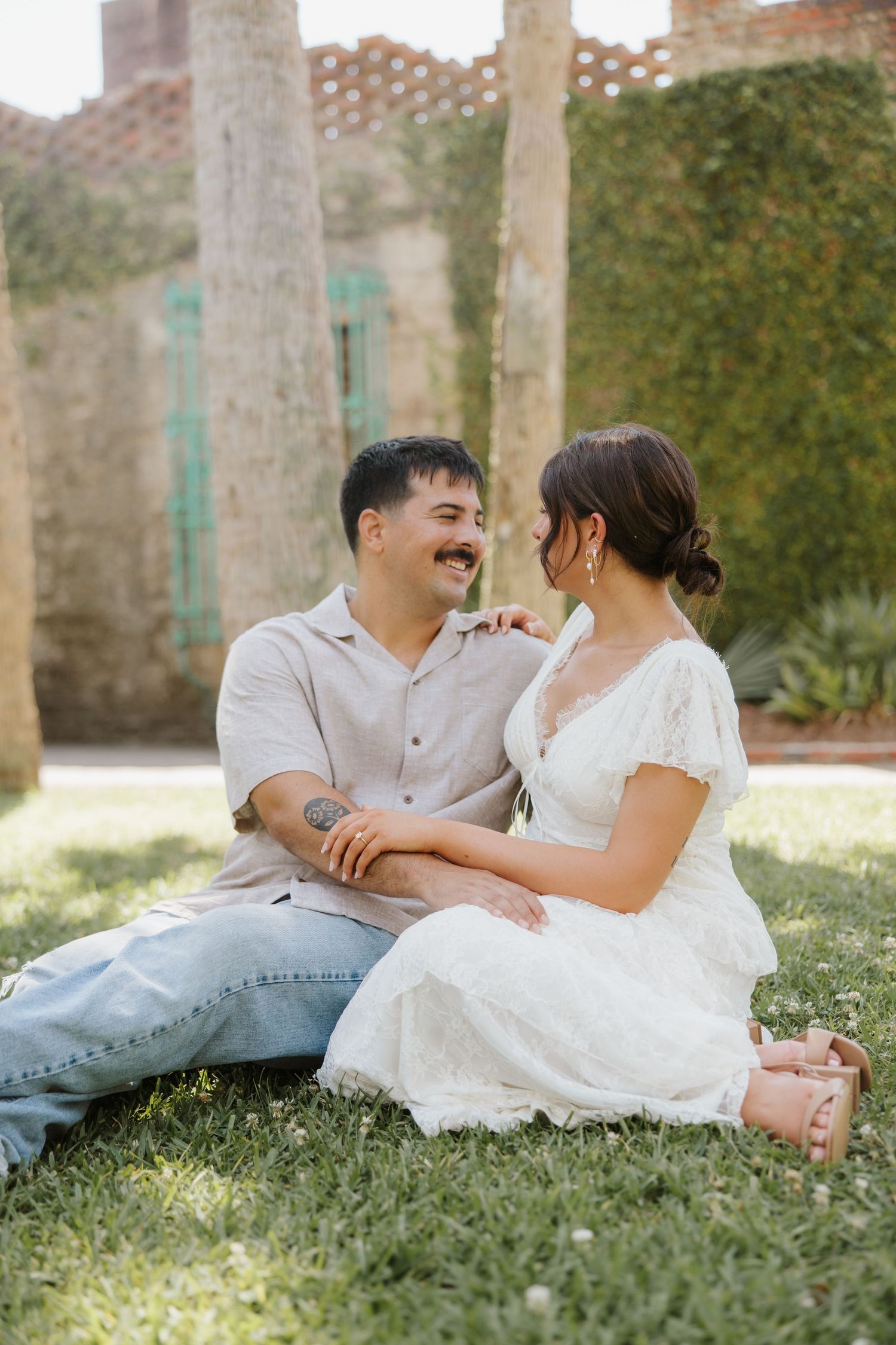 Couple sitting on grass in garden, woman in white lace dress, man in beige shirt and jeans, smiling at each other.