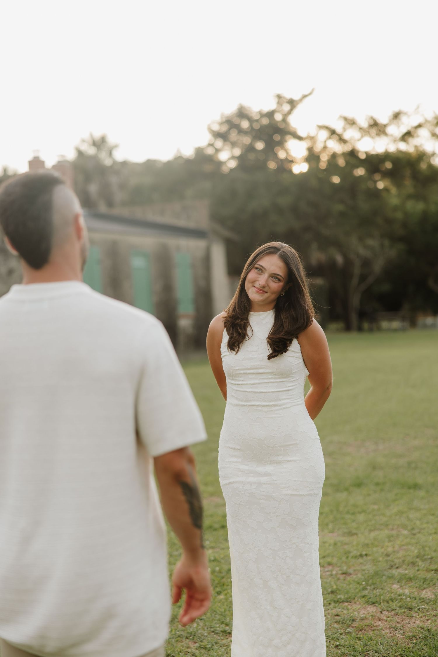 Bride in white fitted gown smiling at groom outdoors at golden hour.