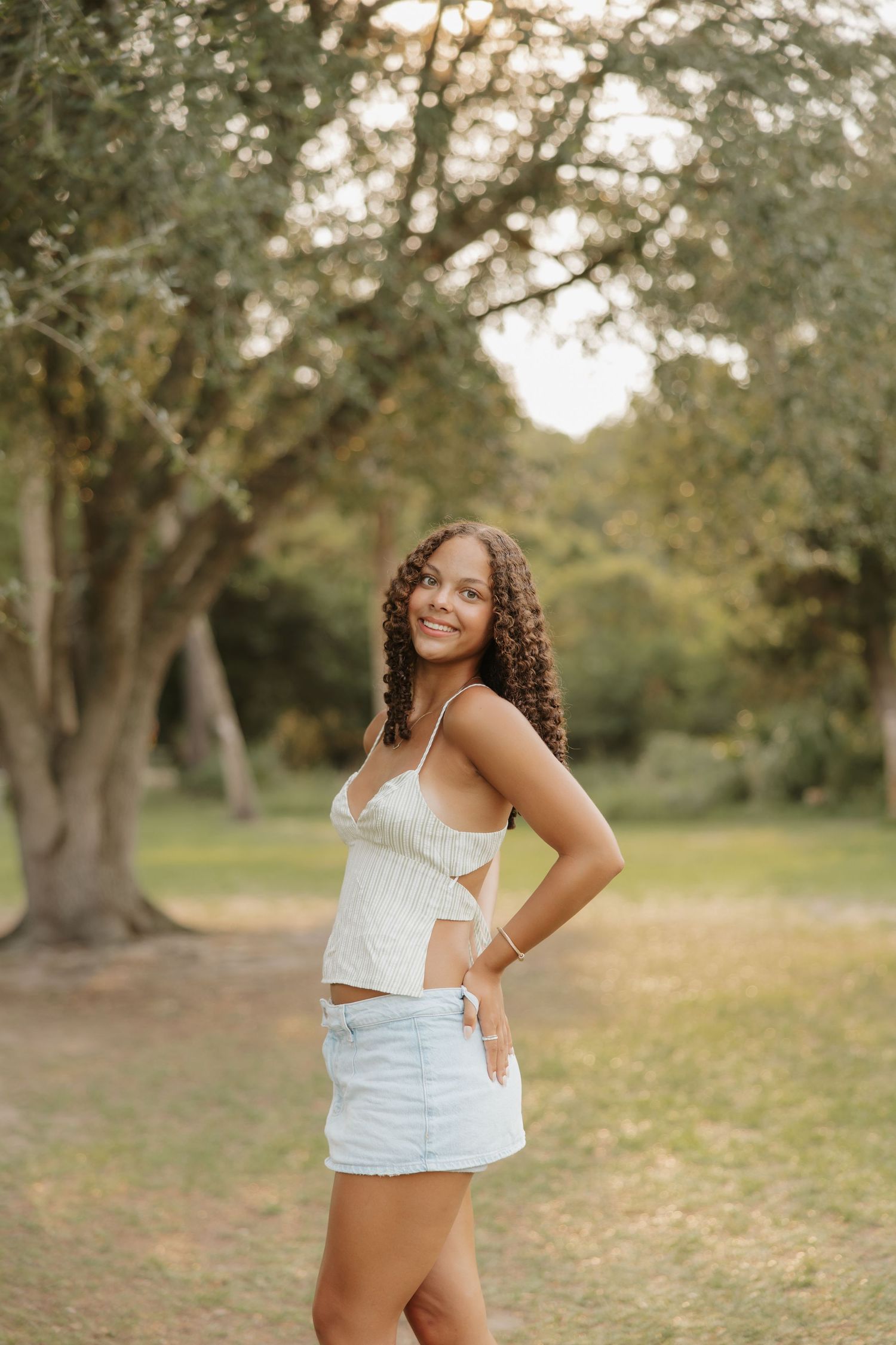 Young woman with curly hair wearing a white crop top and denim skirt posing outdoors in a lush green park.