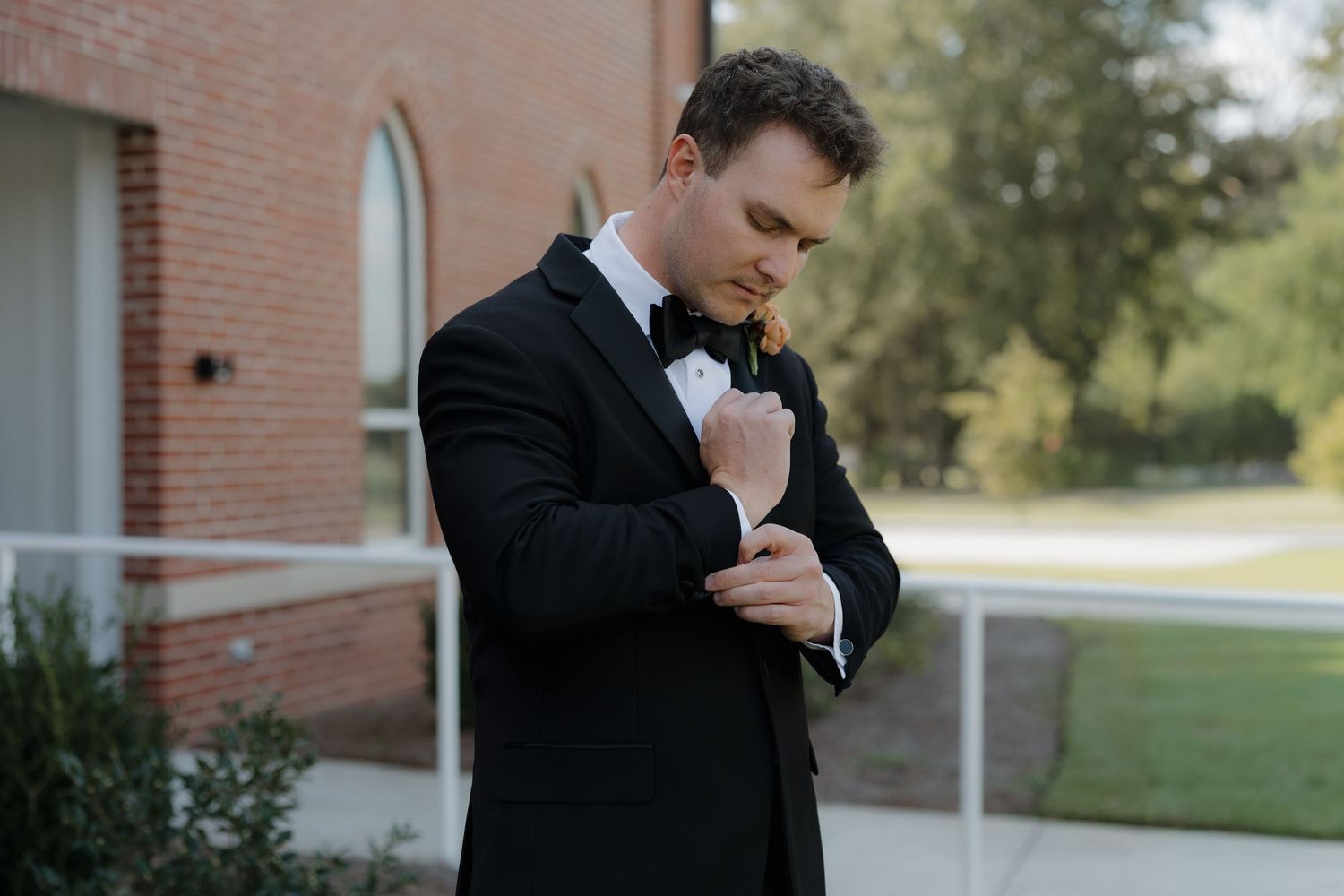 Groom in a black tuxedo and bow tie adjusts his cufflinks outdoors beside a red brick building on a sunny wedding day.