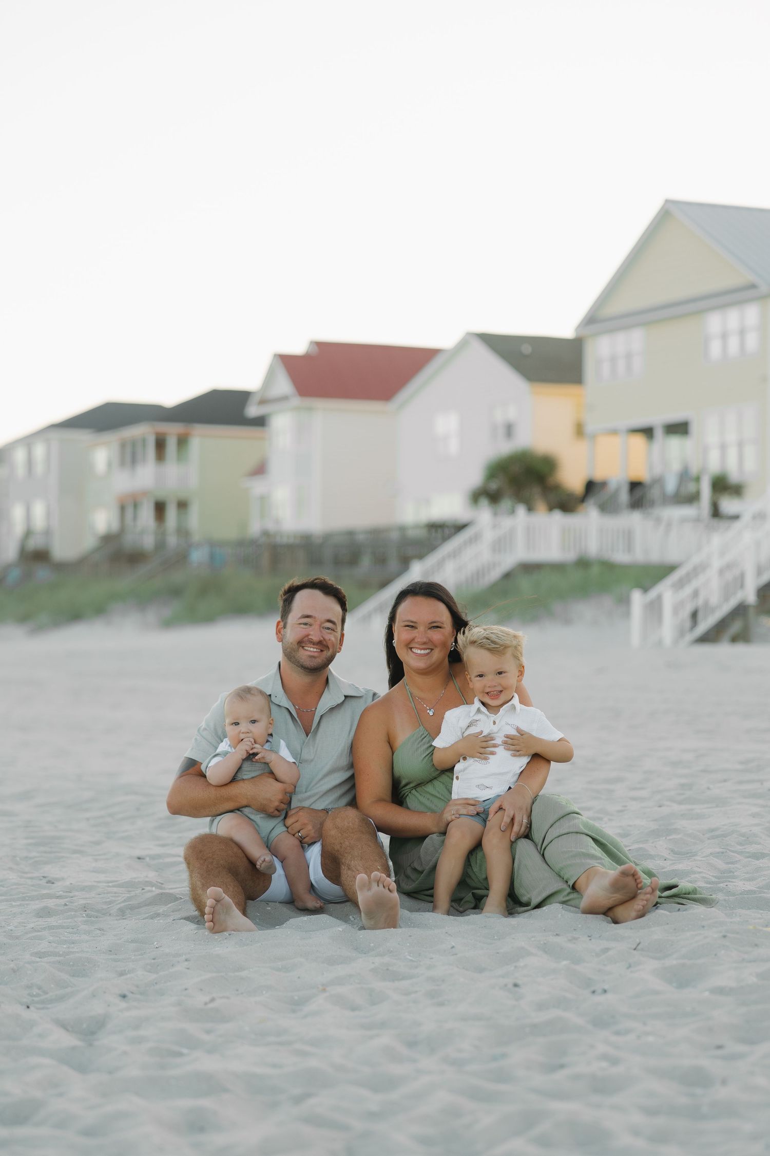 A happy family of four sits on a sandy beach, with colorful beach houses in the background.