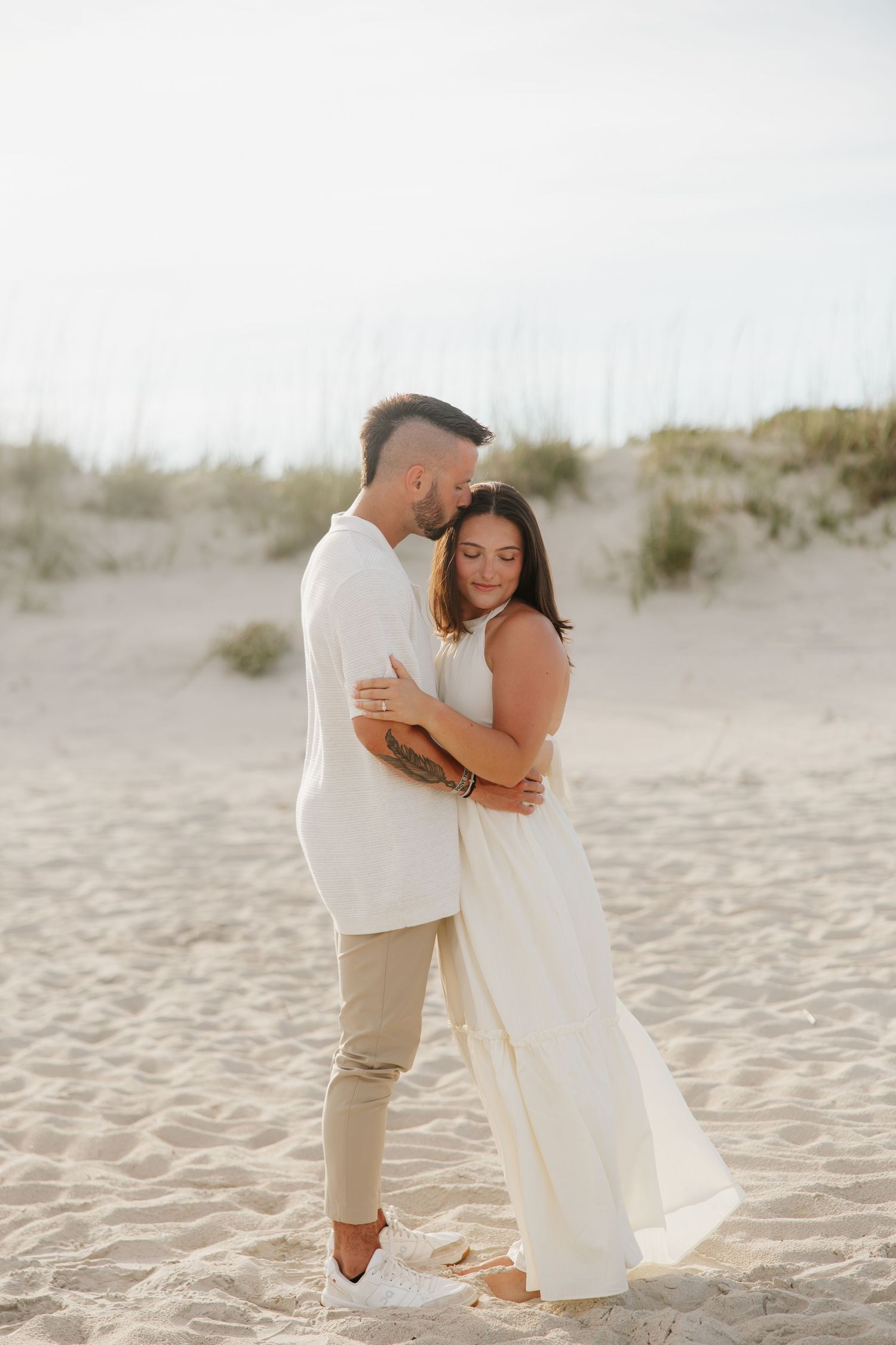 Couple embracing on sandy beach dunes, woman in white dress and man in white shirt and khaki pants.