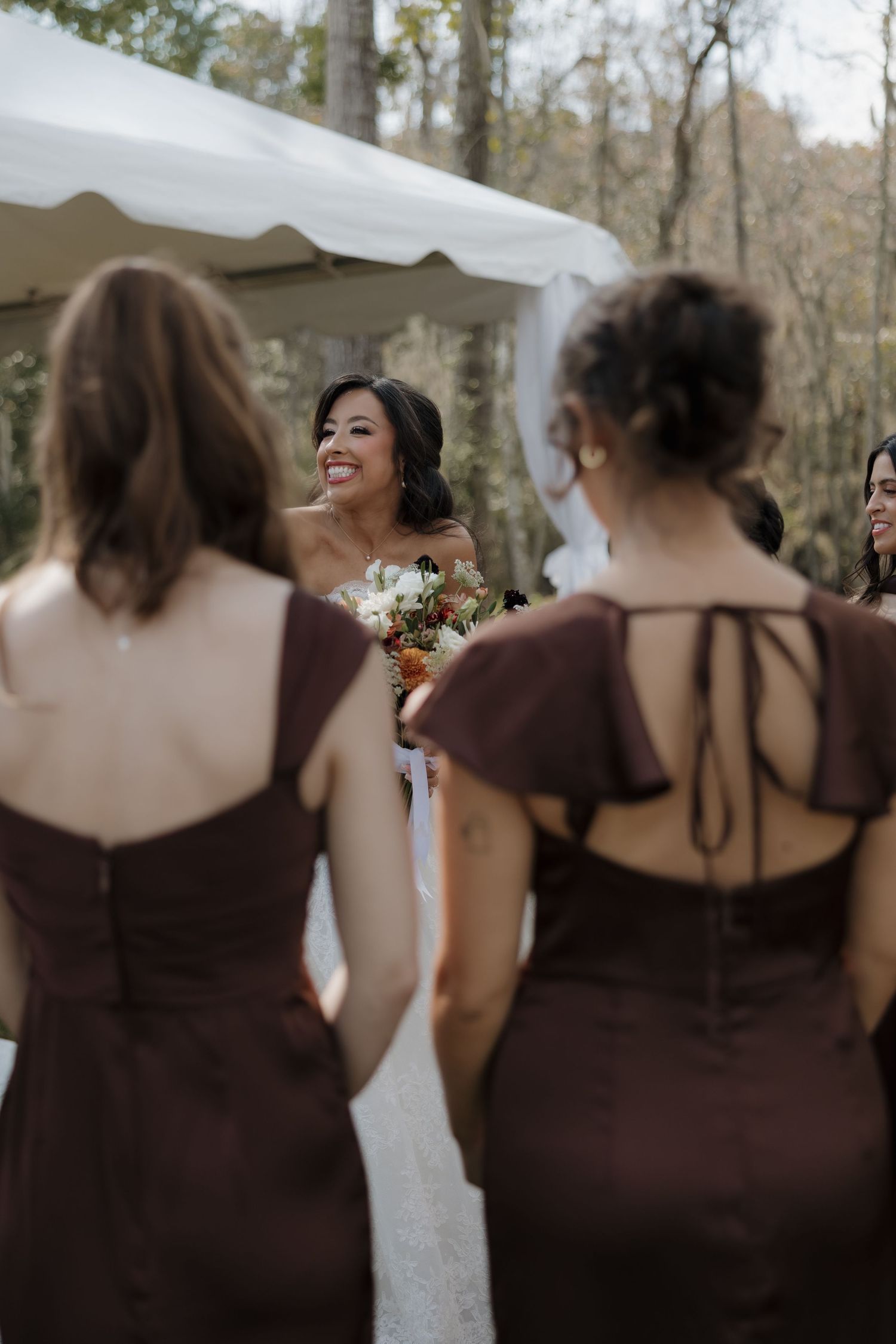 Smiling bride holding a bouquet faces bridesmaids in brown dresses near a white tent at an outdoor woodland wedding.