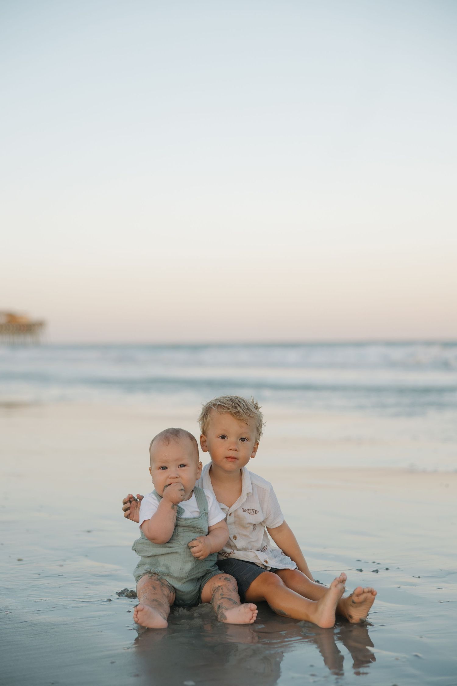 Two young brothers sit together on a sandy beach at sunset, the older boy hugging the baby.