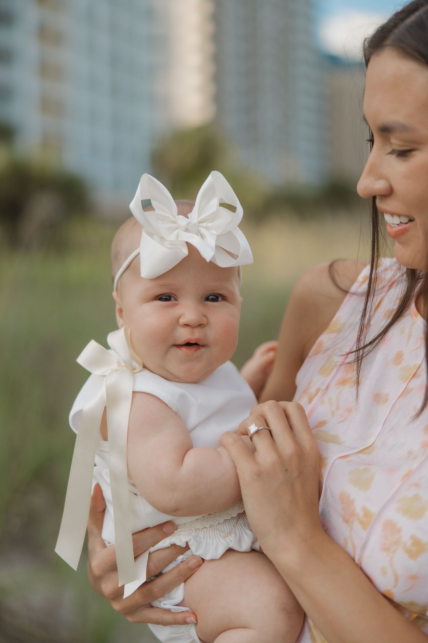 Mother holding adorable baby girl wearing white bow headband and ribbon outfit at beach location.