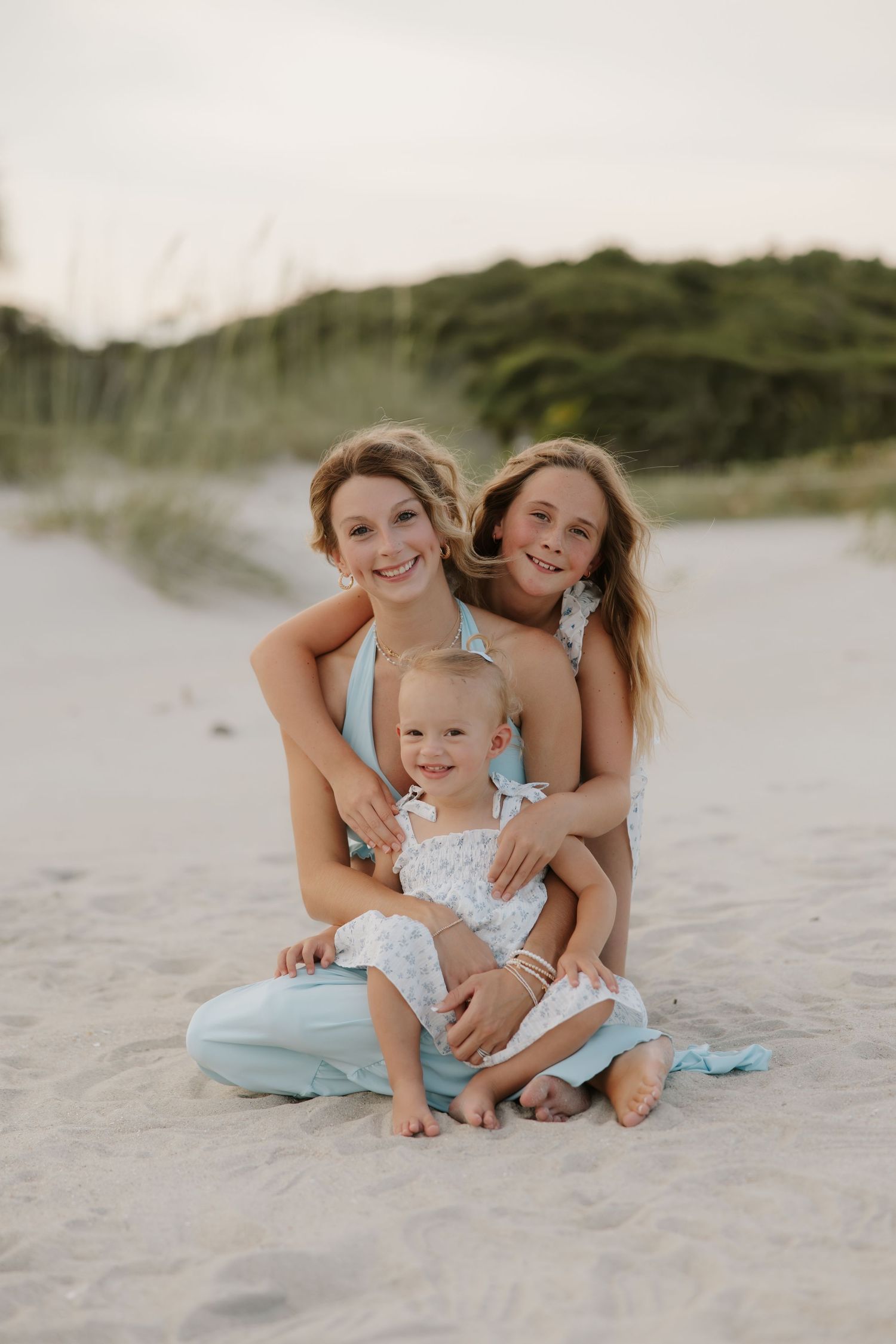 Two sisters sitting on beach sand smiling, holding toddler in white dress at sunset.