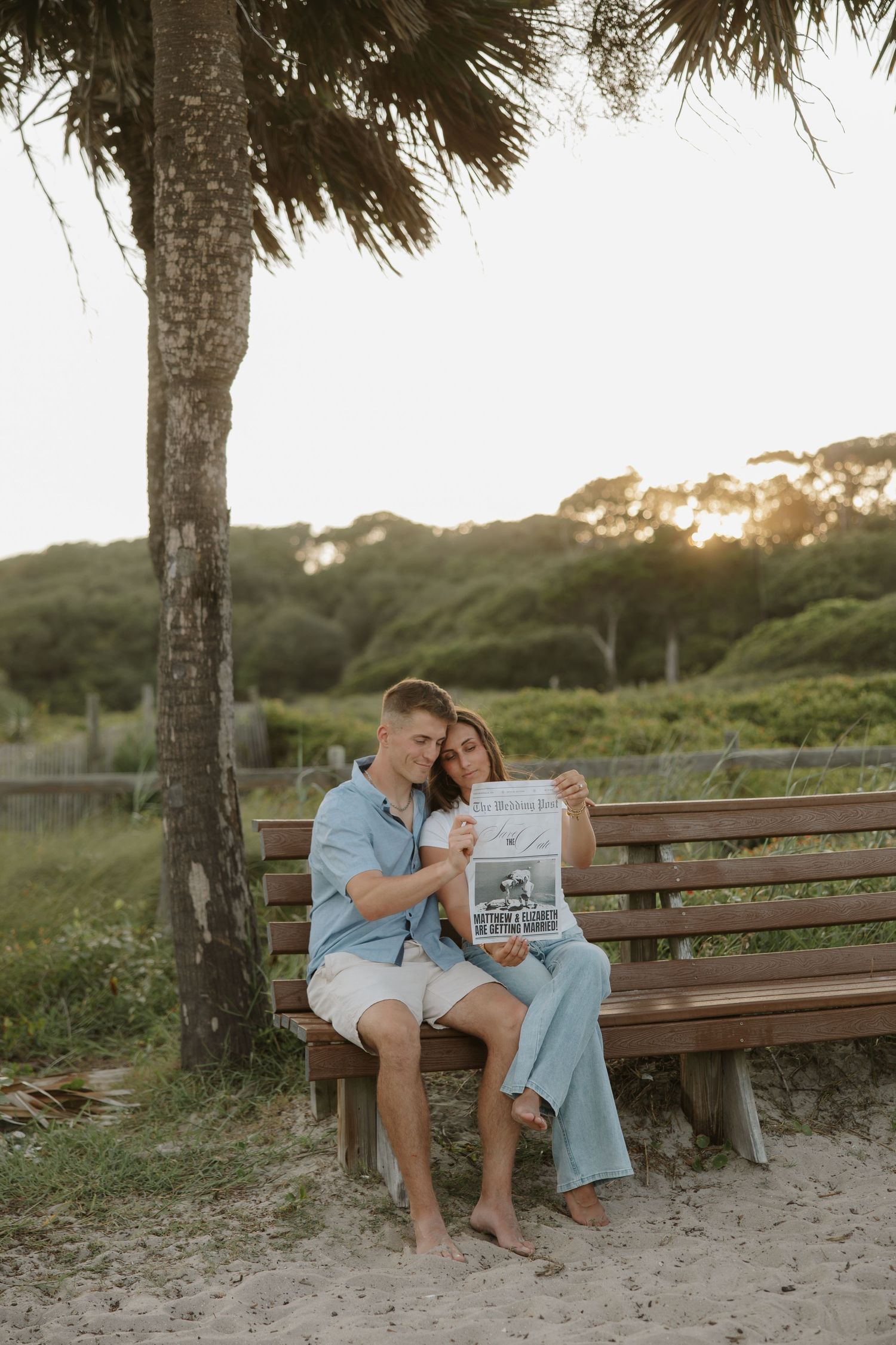 Couple sitting on a wooden bench under a palm tree at sunset, reading a newspaper together near a sandy beach.