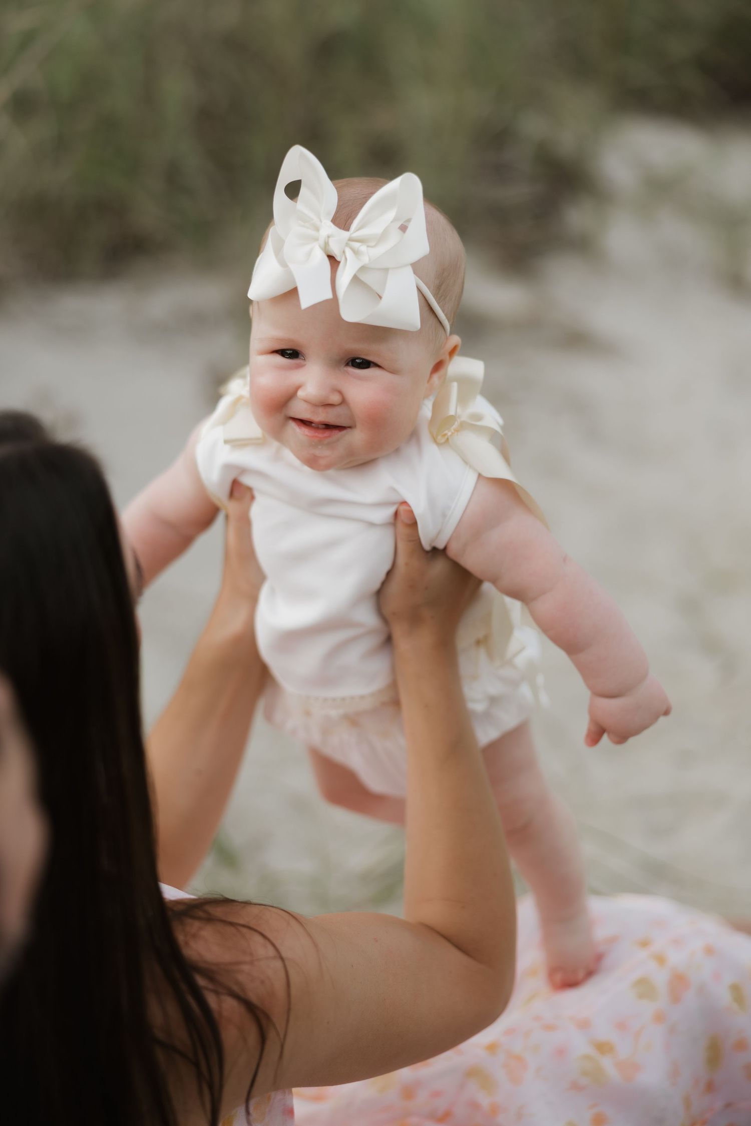 A joyful baby wearing a white bow headband is lifted up by a dark-haired woman on a sandy beach.