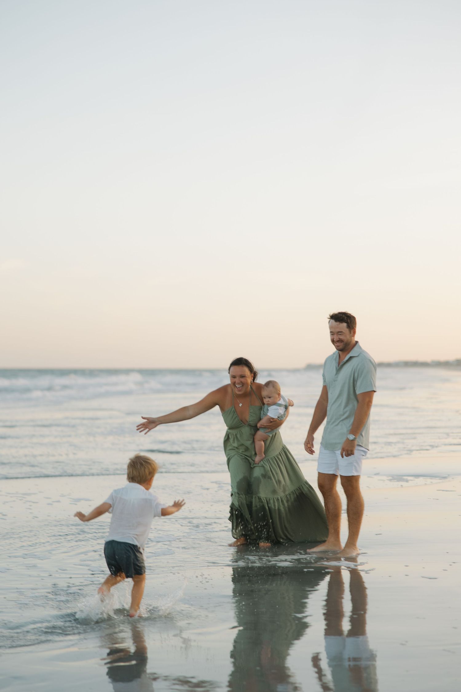 Family enjoying a beach walk at sunset, mother in green dress holding baby while toddler runs in the waves.