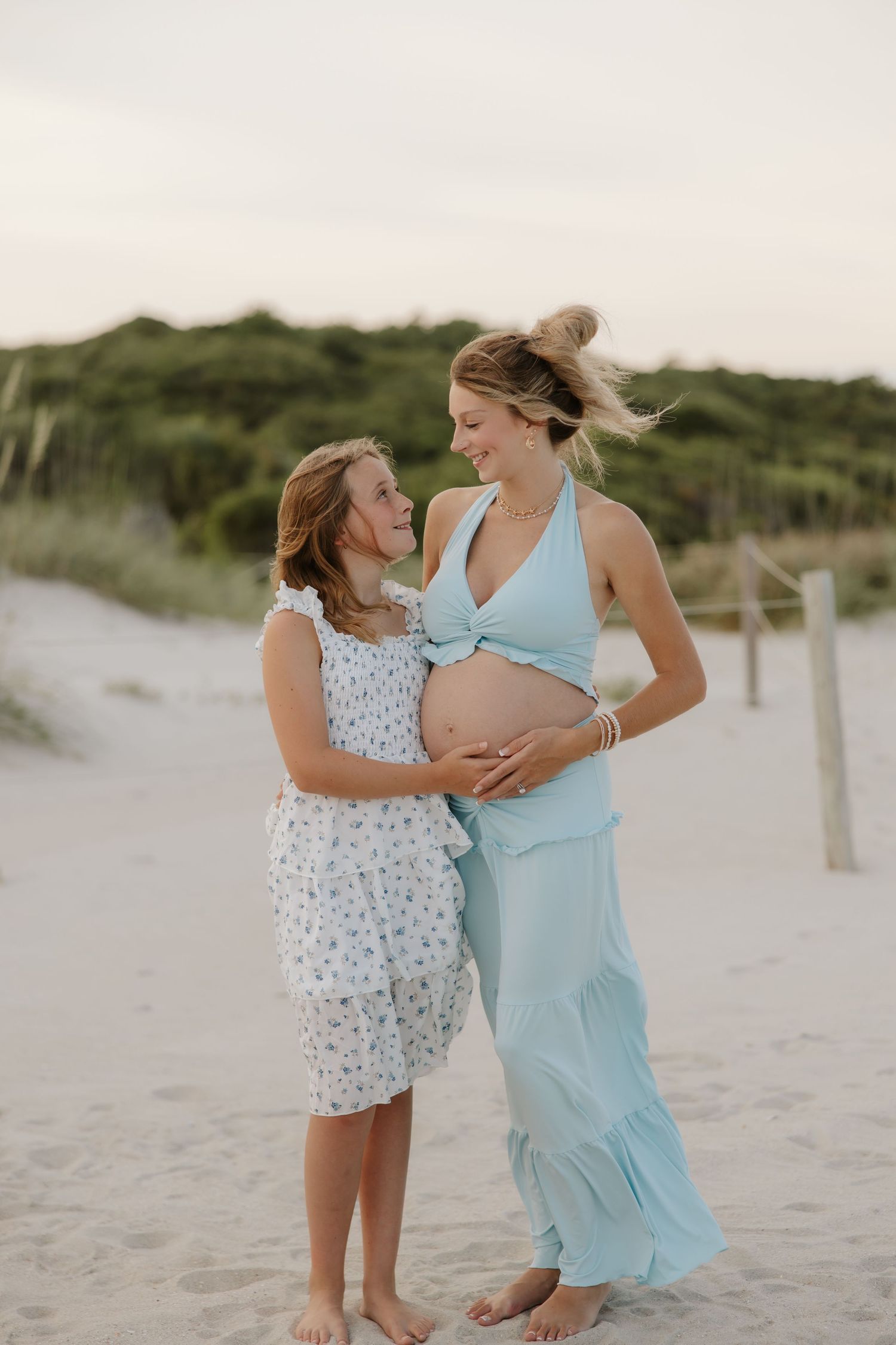 Pregnant woman in light blue dress with young girl touching her baby bump on sandy beach at dusk.