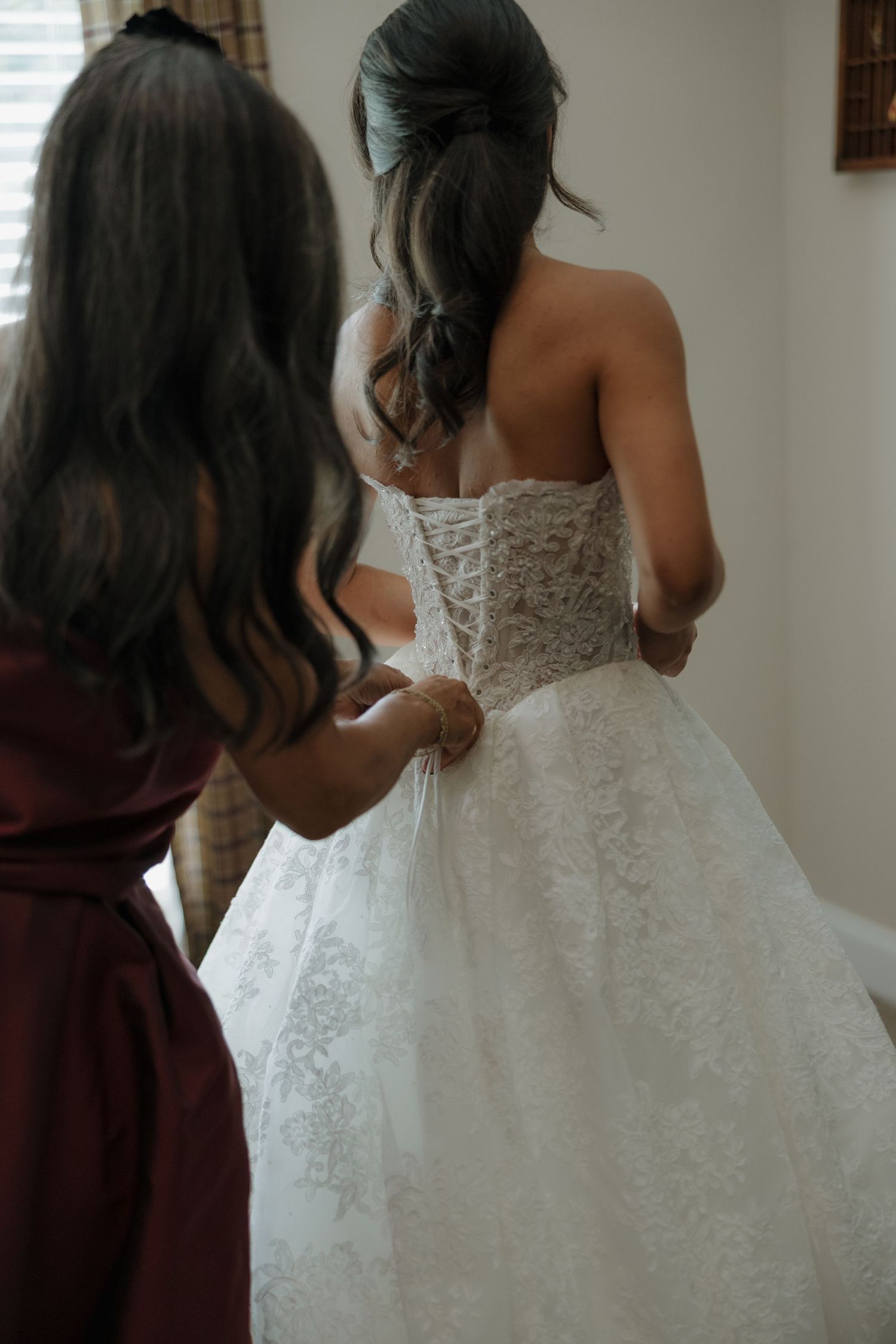 A bridesmaid in a burgundy dress laces up the corset back of a bride's strapless white lace wedding gown.