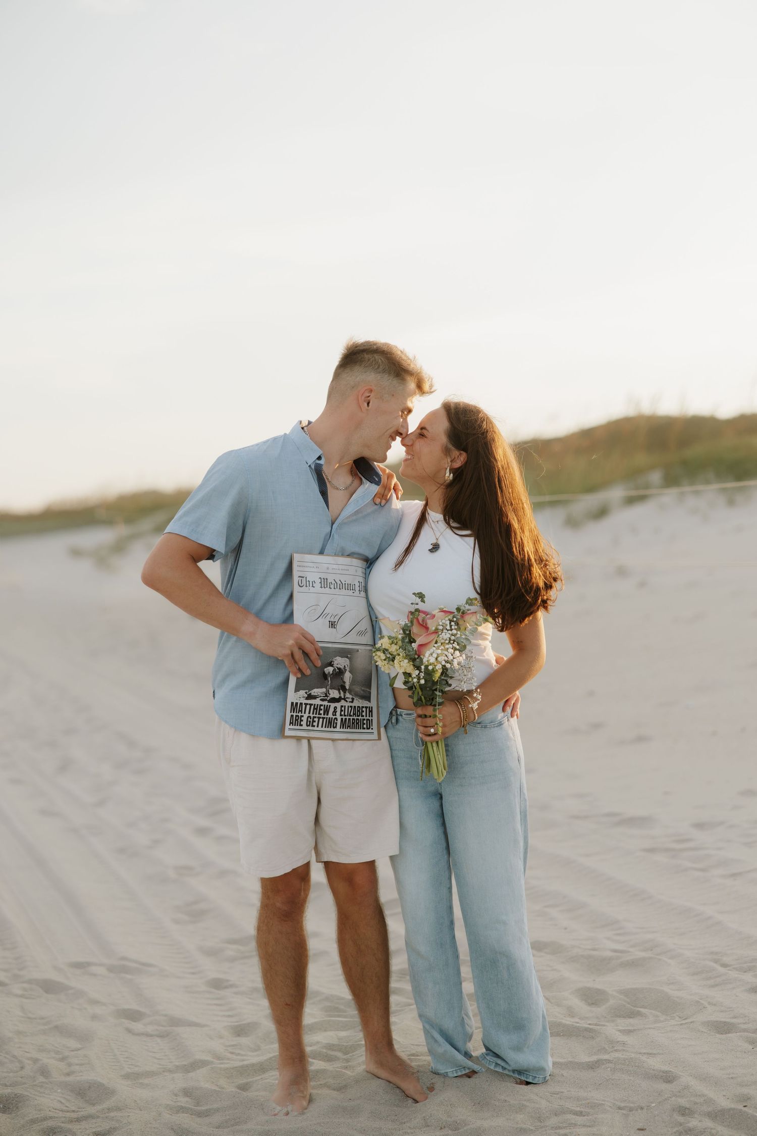 Couple sharing a romantic moment on a sandy beach, holding flowers and a newspaper at sunset.