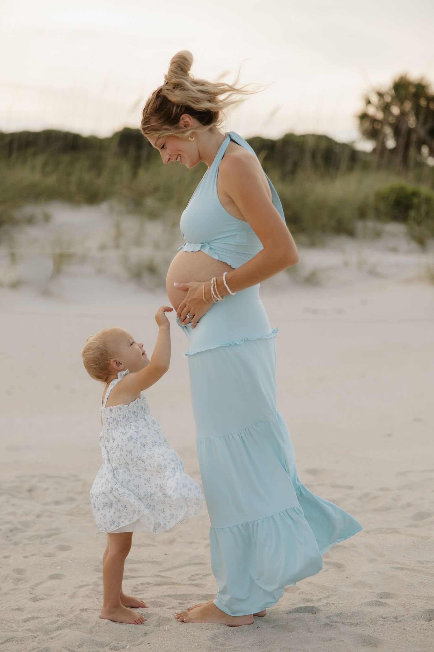 Pregnant woman in light blue dress on beach with toddler touching her baby bump at sunset.